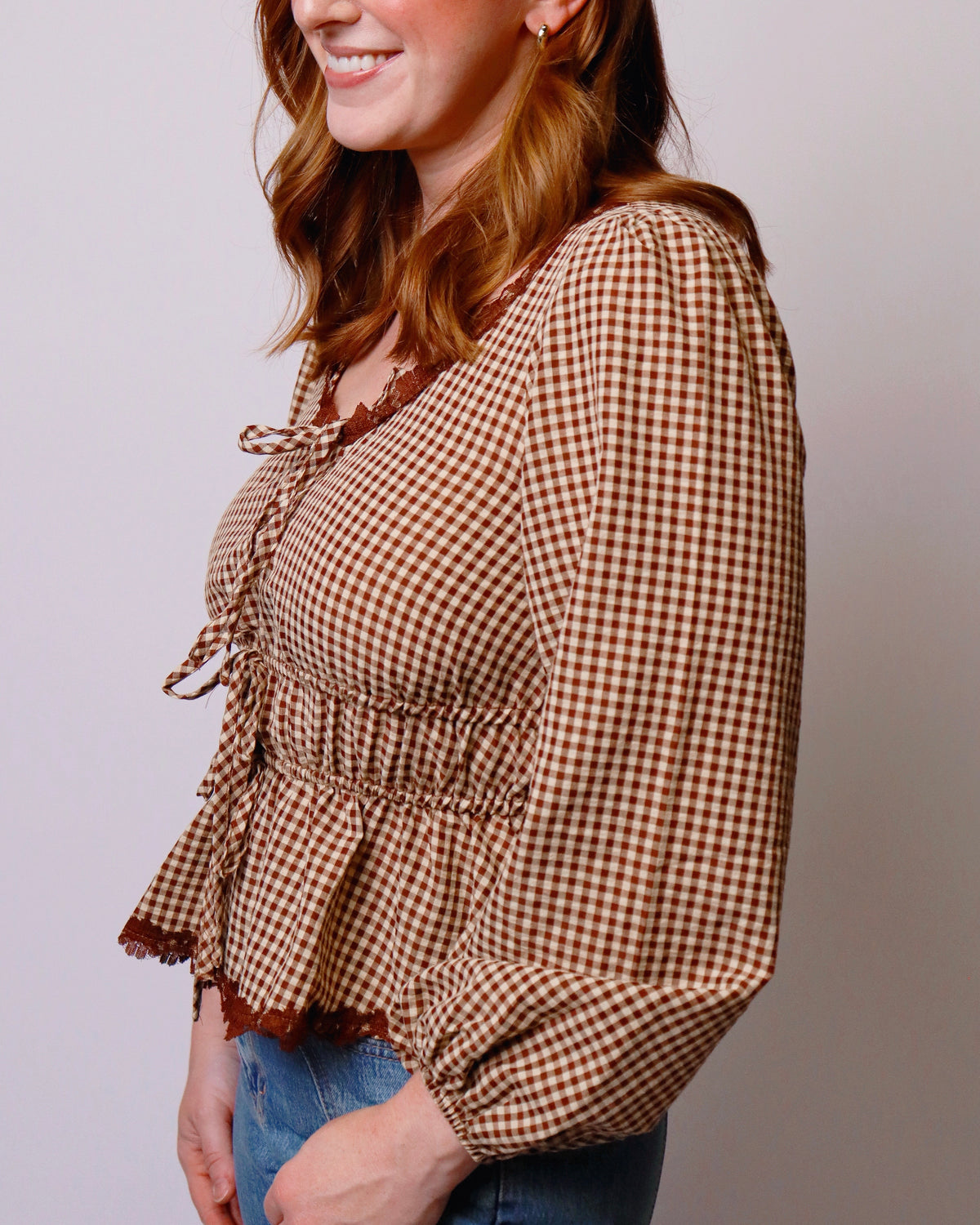 A woman wearing a brown and beige gingham long-sleeve blouse with a smocked waist and tie detail at the neckline, paired with blue jeans against a plain light gray background.