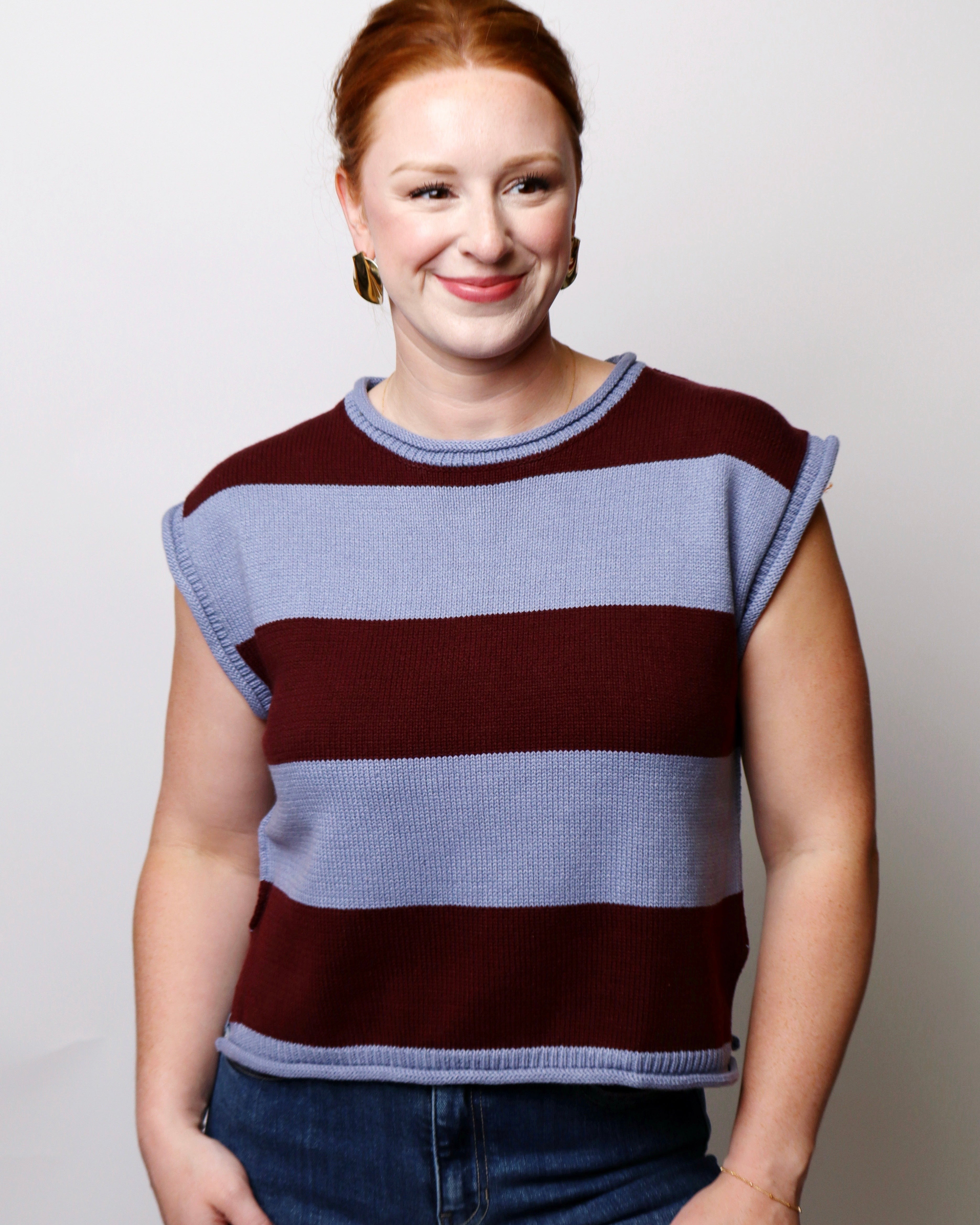 A woman with red hair tied back, wearing a sleeveless knit top with horizontal stripes in maroon and light blue, paired with blue jeans, standing against a plain light gray background.