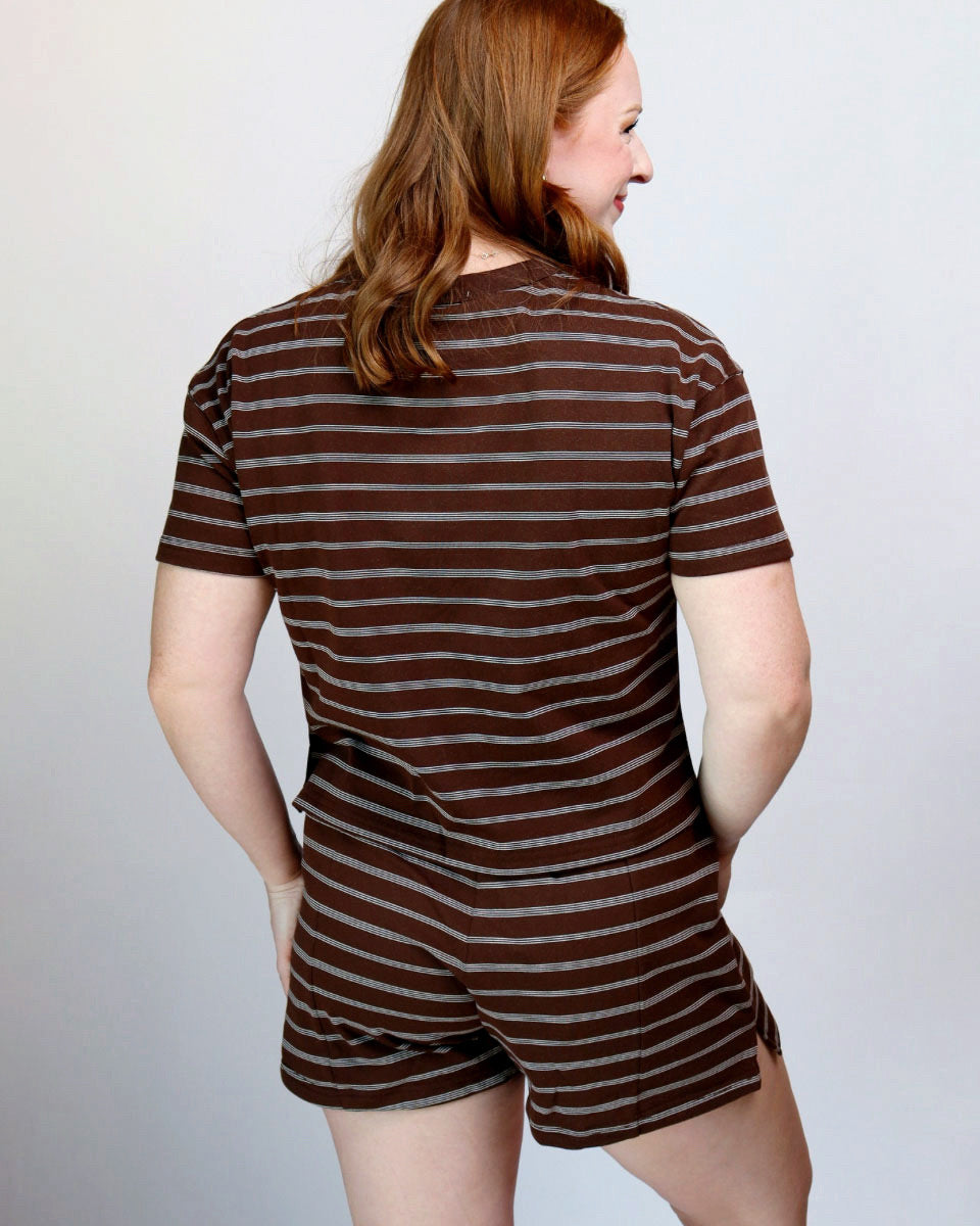 A woman with red hair is shown from the back wearing a brown and gray striped short-sleeve romper against a light gray studio background.