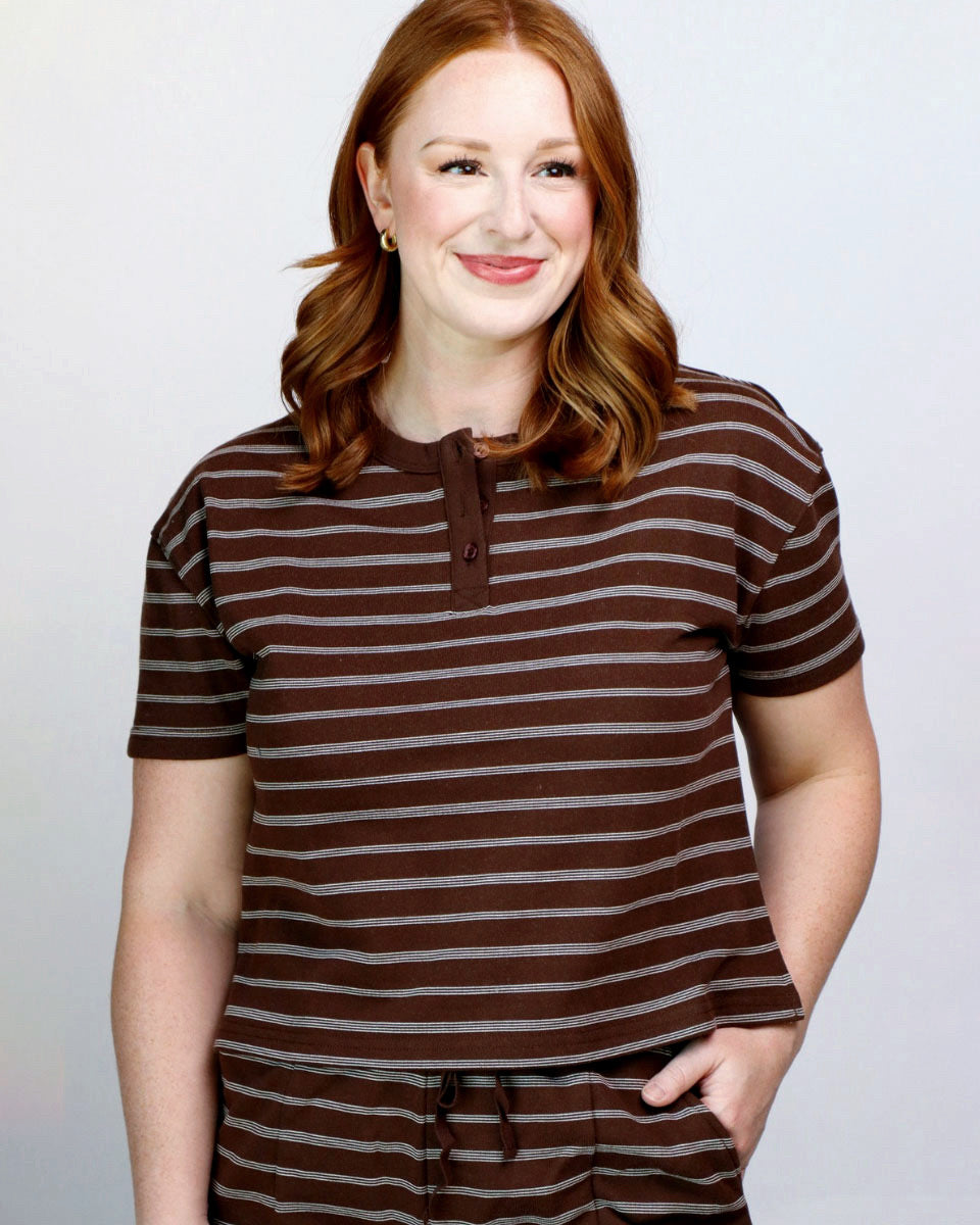 A woman with red hair wearing a brown and gray striped short-sleeve henley top and matching striped pants, standing against a light gray studio background.