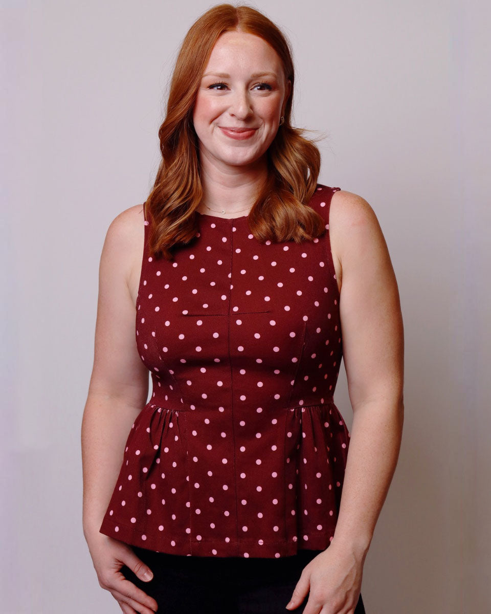 A woman with red hair wearing a sleeveless burgundy top with white polka dots, standing against a light gray studio background.
