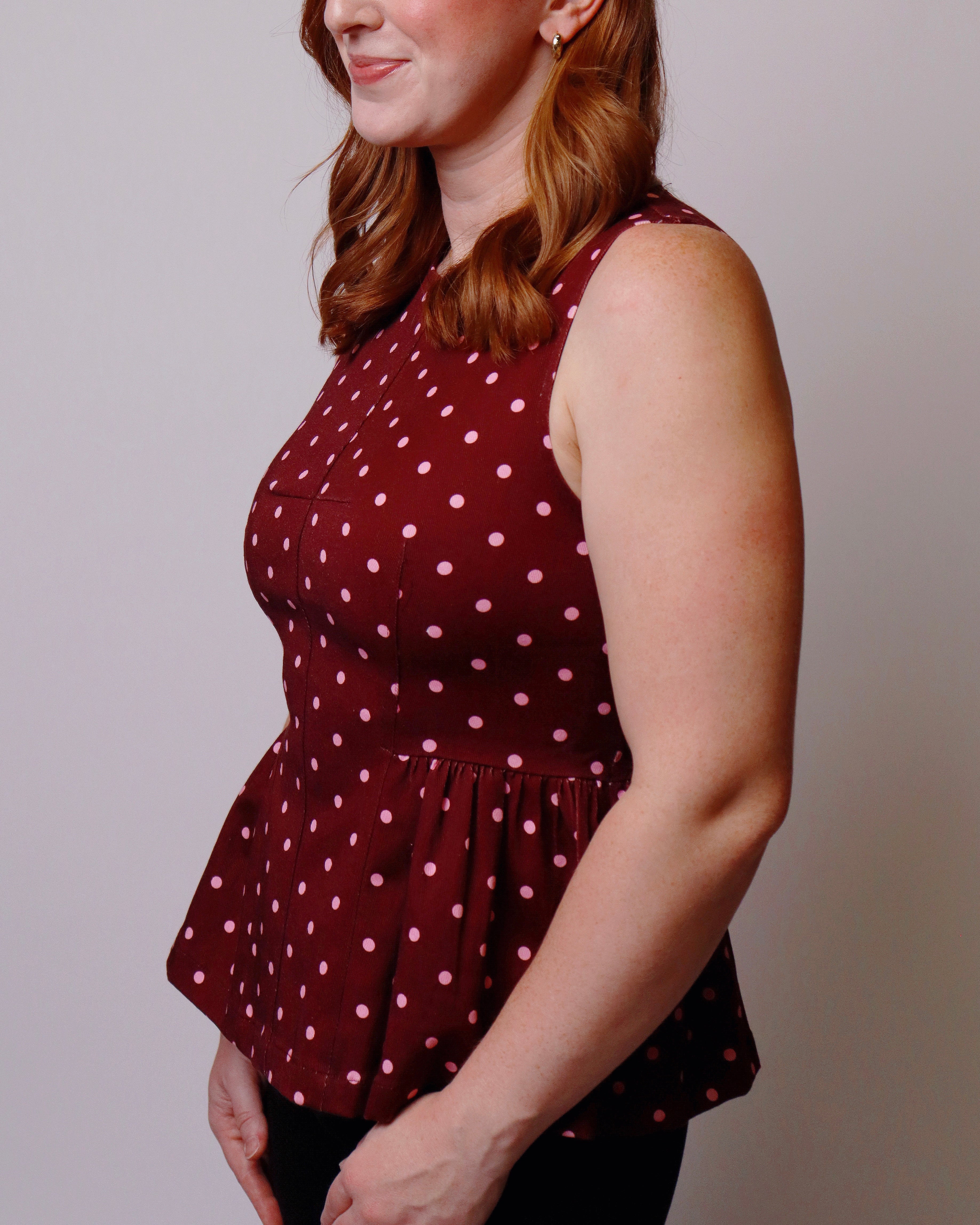 A woman wearing a sleeveless peplum top in burgundy with a pink polka dot pattern, shown from a side profile against a plain light gray background.