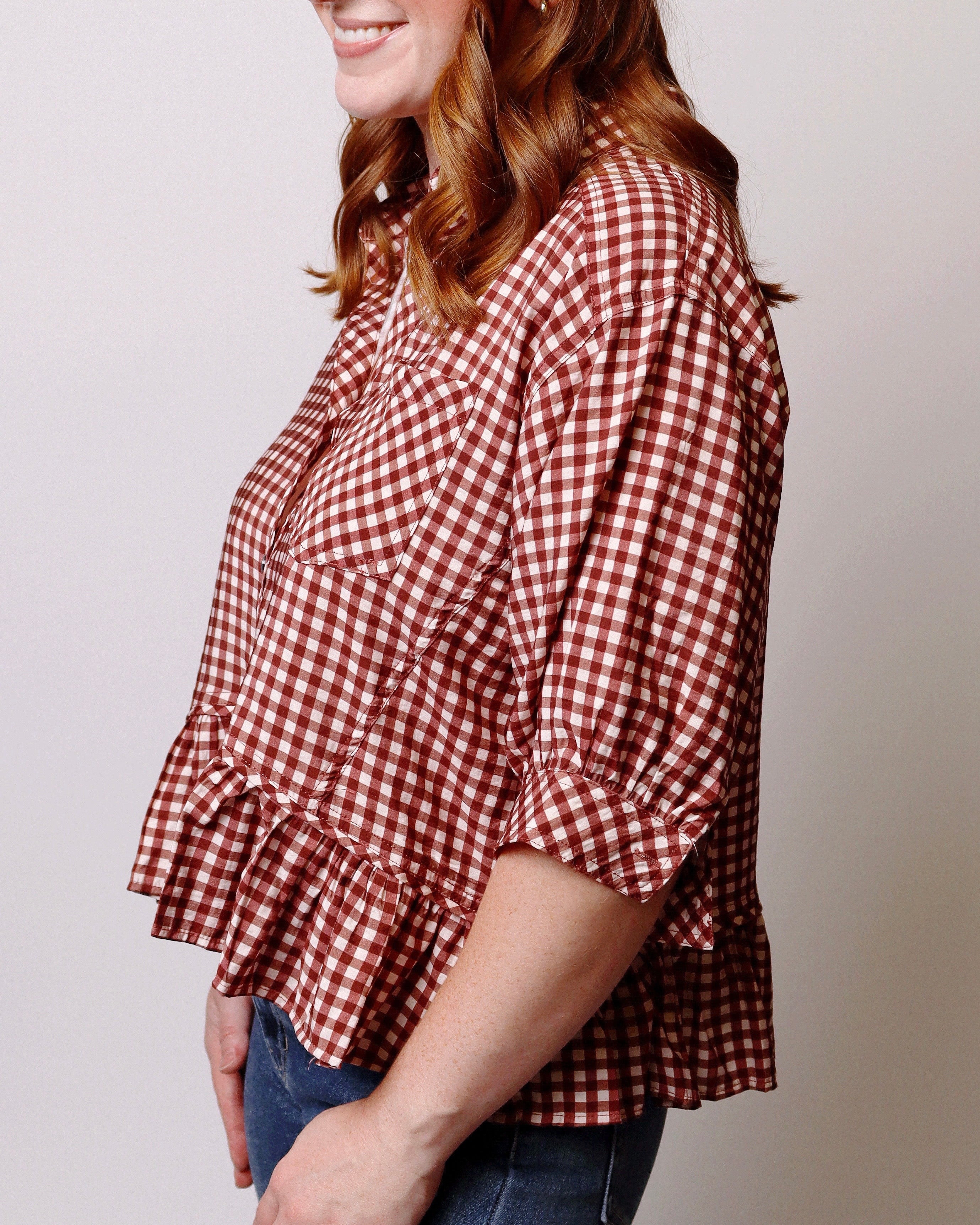 A woman wearing a red and white gingham check blouse with a ruffled peplum hem and elbow-length sleeves, paired with blue jeans, shown from a side profile against a plain light gray background.