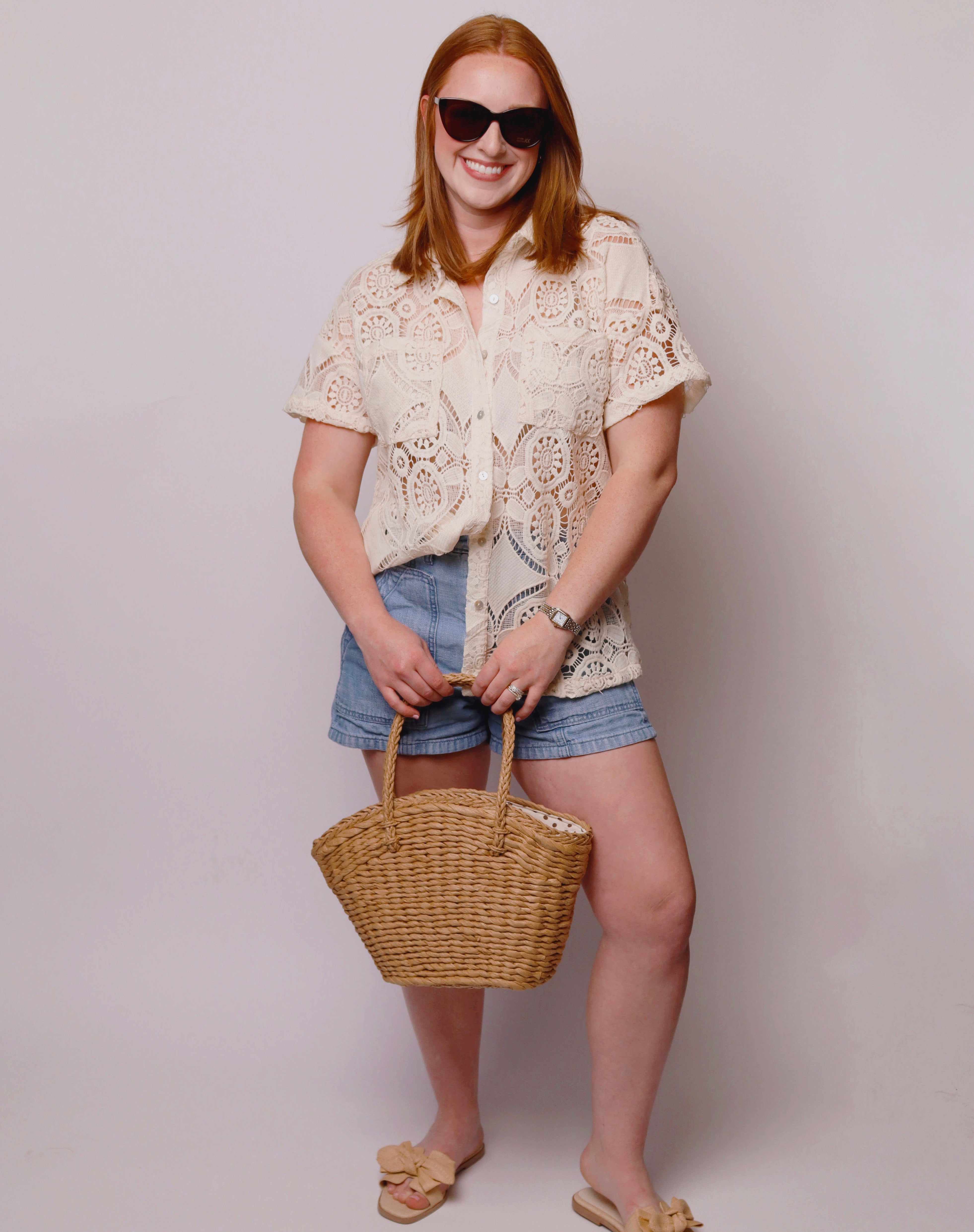 Woman wearing a white lace top and denim shorts, holding a wicker bag against a plain background