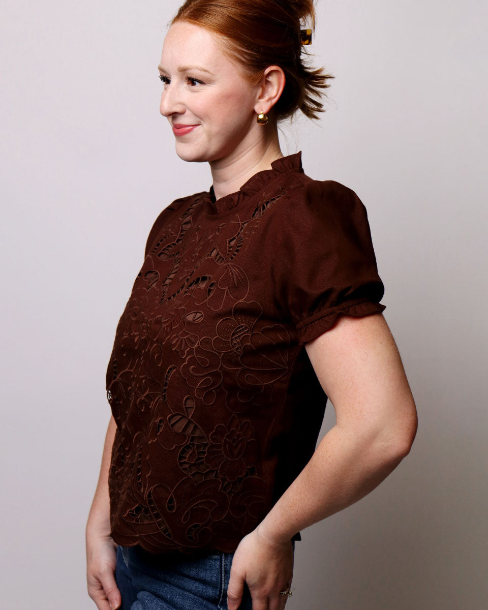 A woman with red hair tied back is wearing a dark brown short-sleeve blouse with tonal floral embroidery, paired with blue jeans, against a plain light gray background.