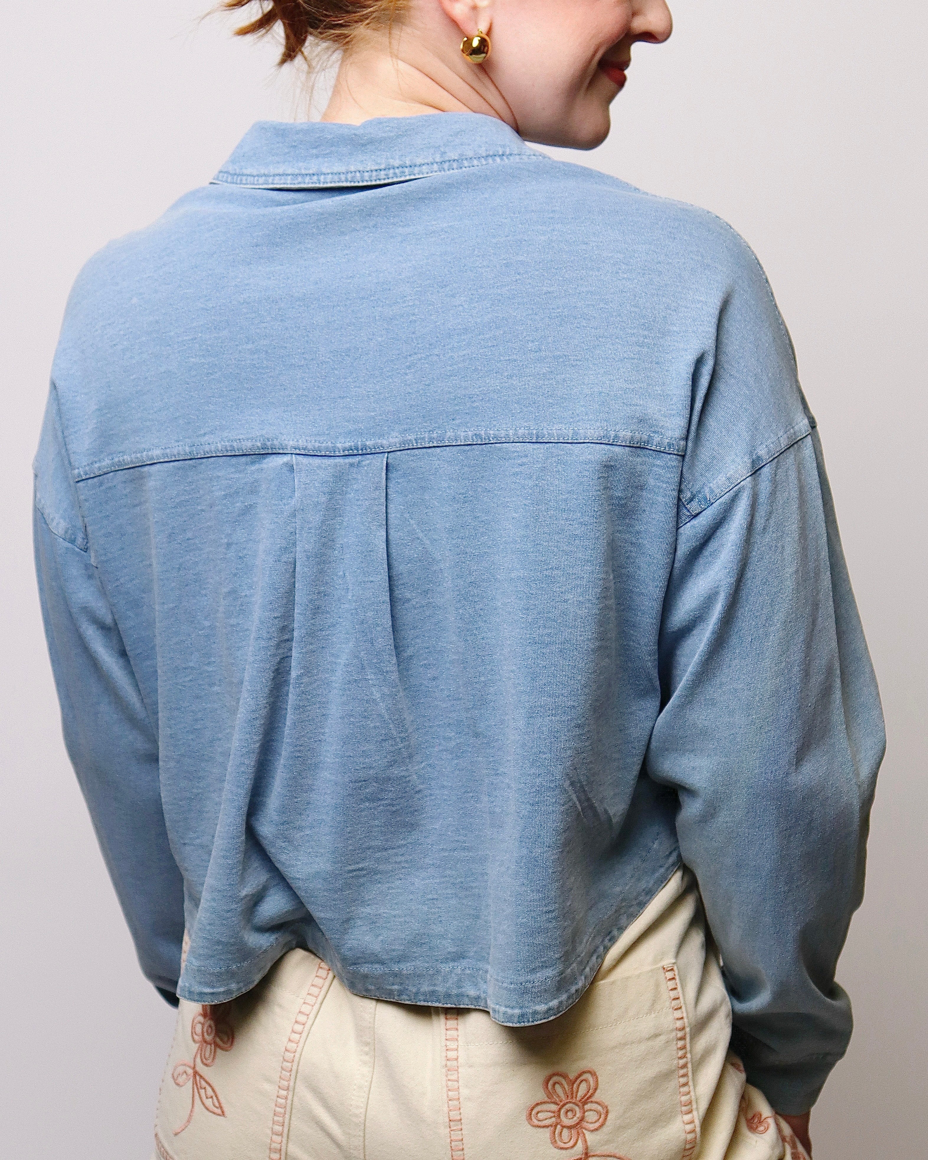 A person shown from the back wearing a light blue cropped button-up shirt and cream-colored pants with pink floral embroidery, against a plain light gray background.