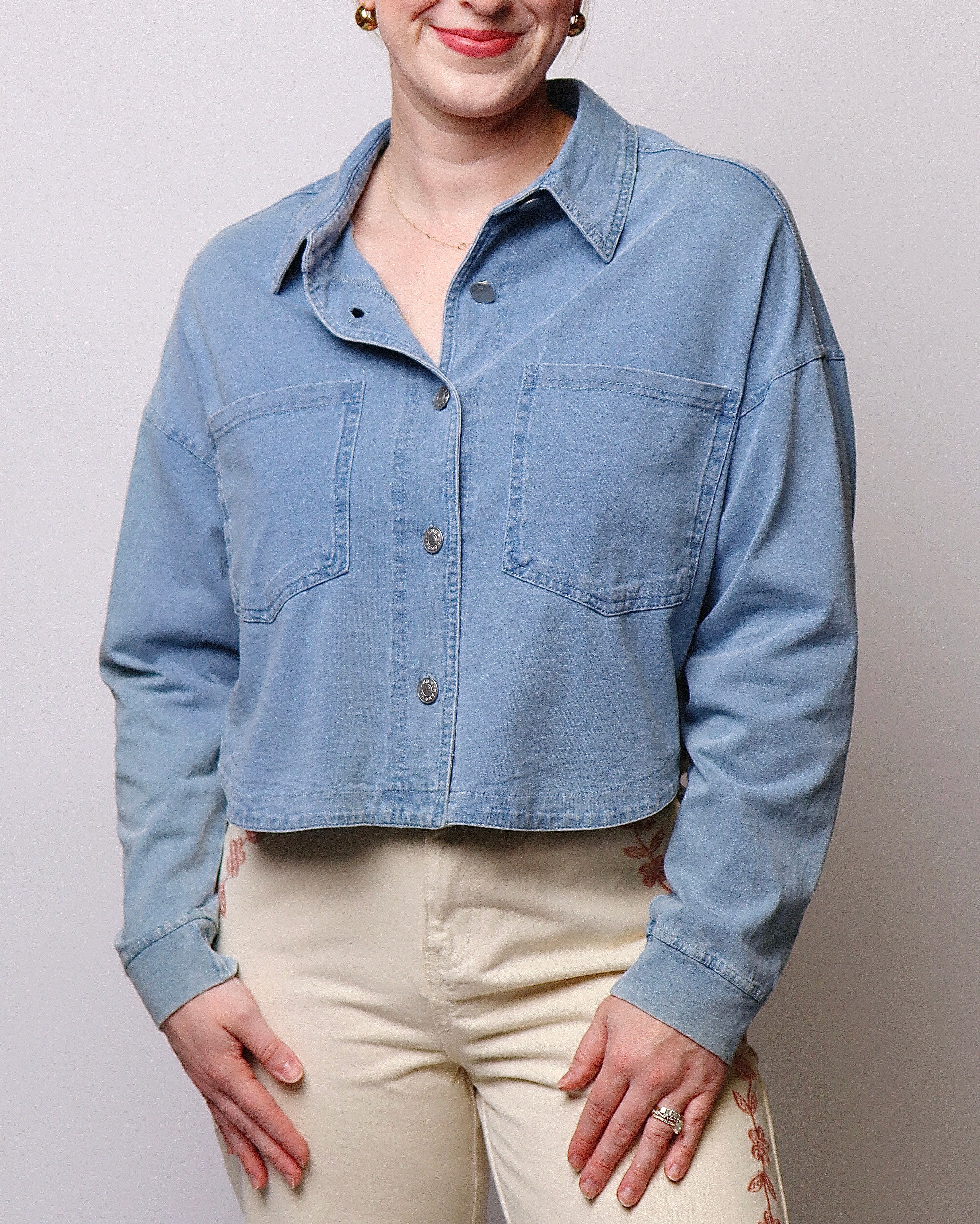 A woman wearing a cropped light blue denim shirt-jacket with front chest pockets and cream-colored pants, shown from the mouth down against a plain light gray studio background.