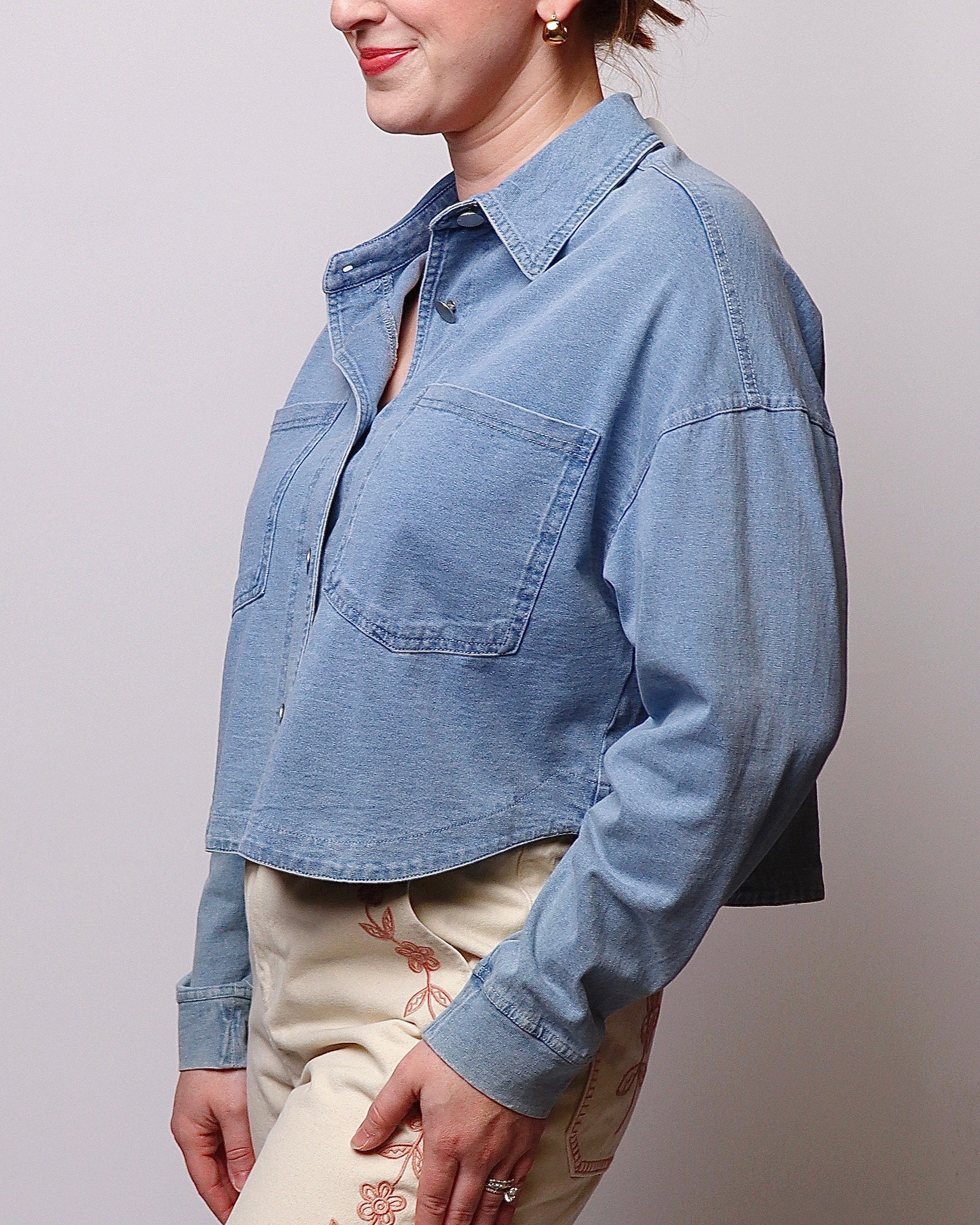 A woman wearing a cropped light blue denim shirt jacket with chest pockets, paired with cream floral pants, shown from the side against a plain light gray background.