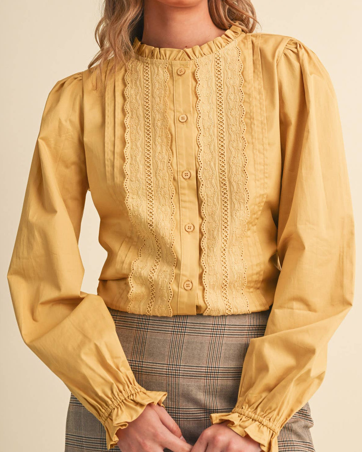 A woman wearing a honey gold long sleeve button up blouse with lace details on a neutral background 