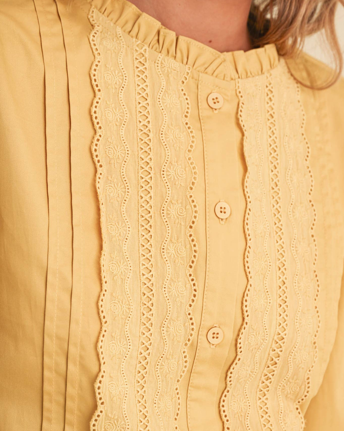A woman wearing a honey gold long sleeve button up blouse with lace details on a neutral background close up