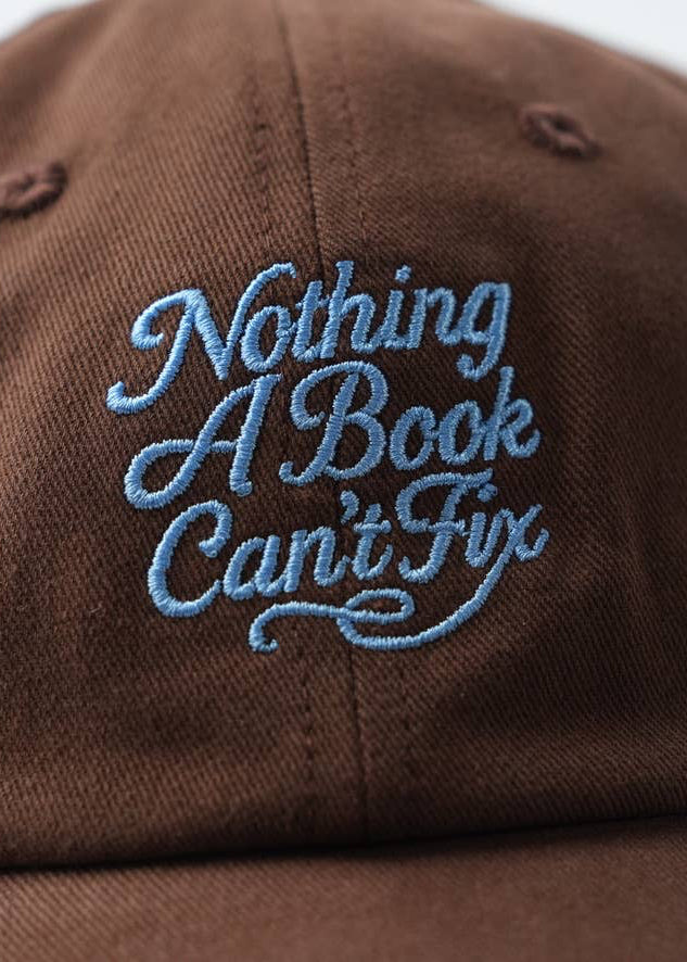Brown cap with blue embroidered text 'Nothing A Book Can't Fix' on a white background