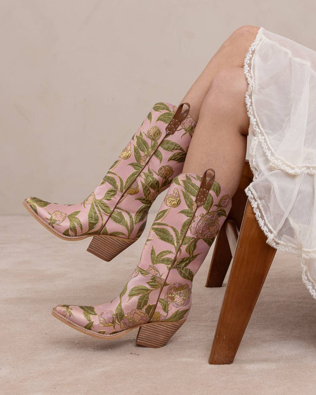 Floral embroidered cowboy boots worn with a white lace skirt on a neutral background