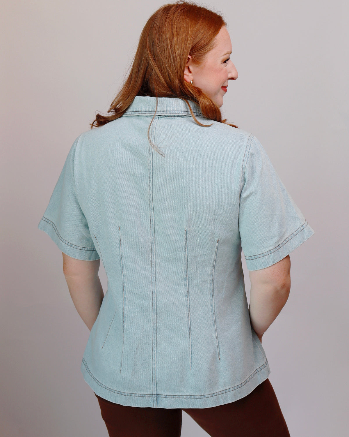 A woman with red hair is shown from the back wearing a light blue denim short-sleeve shirt with vertical seam detailing on the back, paired with brown pants, against a plain light gray background.