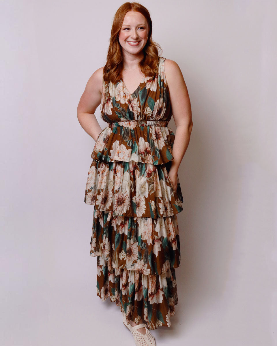 A woman with red hair wearing a sleeveless, tiered maxi dress with a brown, beige, and green floral pattern, standing against a light gray studio background.
