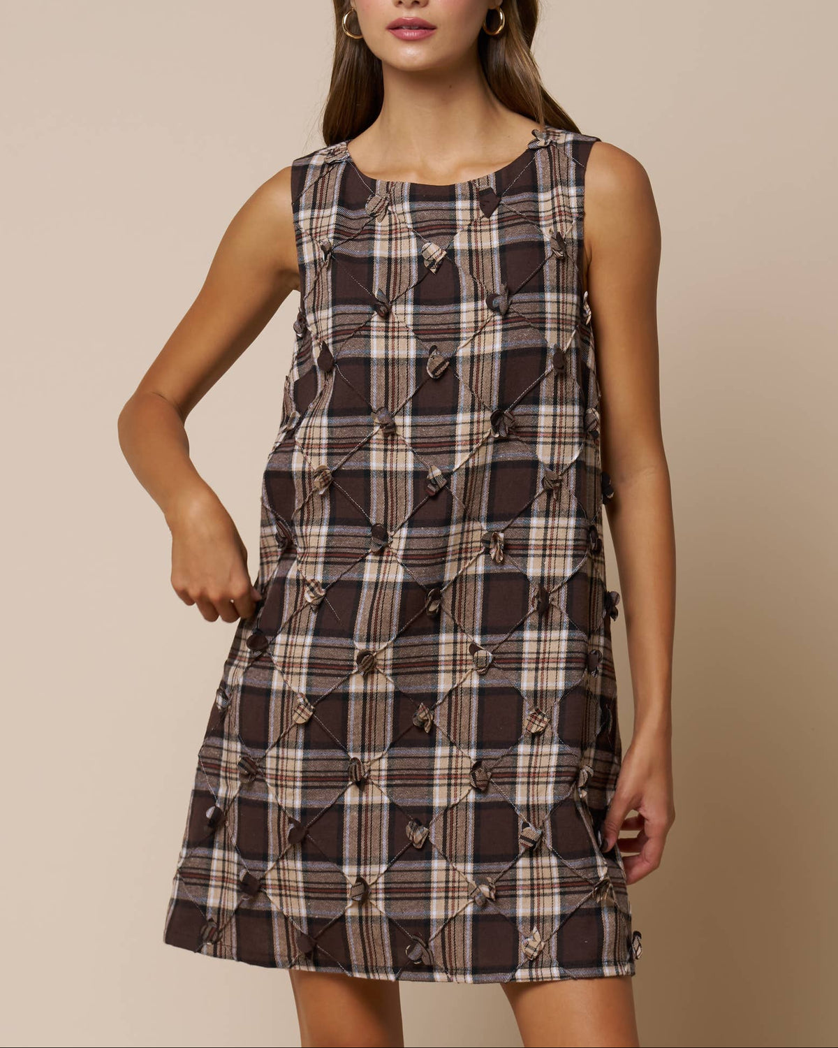 Model weaing a brown/taupe plaid sleeveless mini dress with diamond flower details on a neutral background