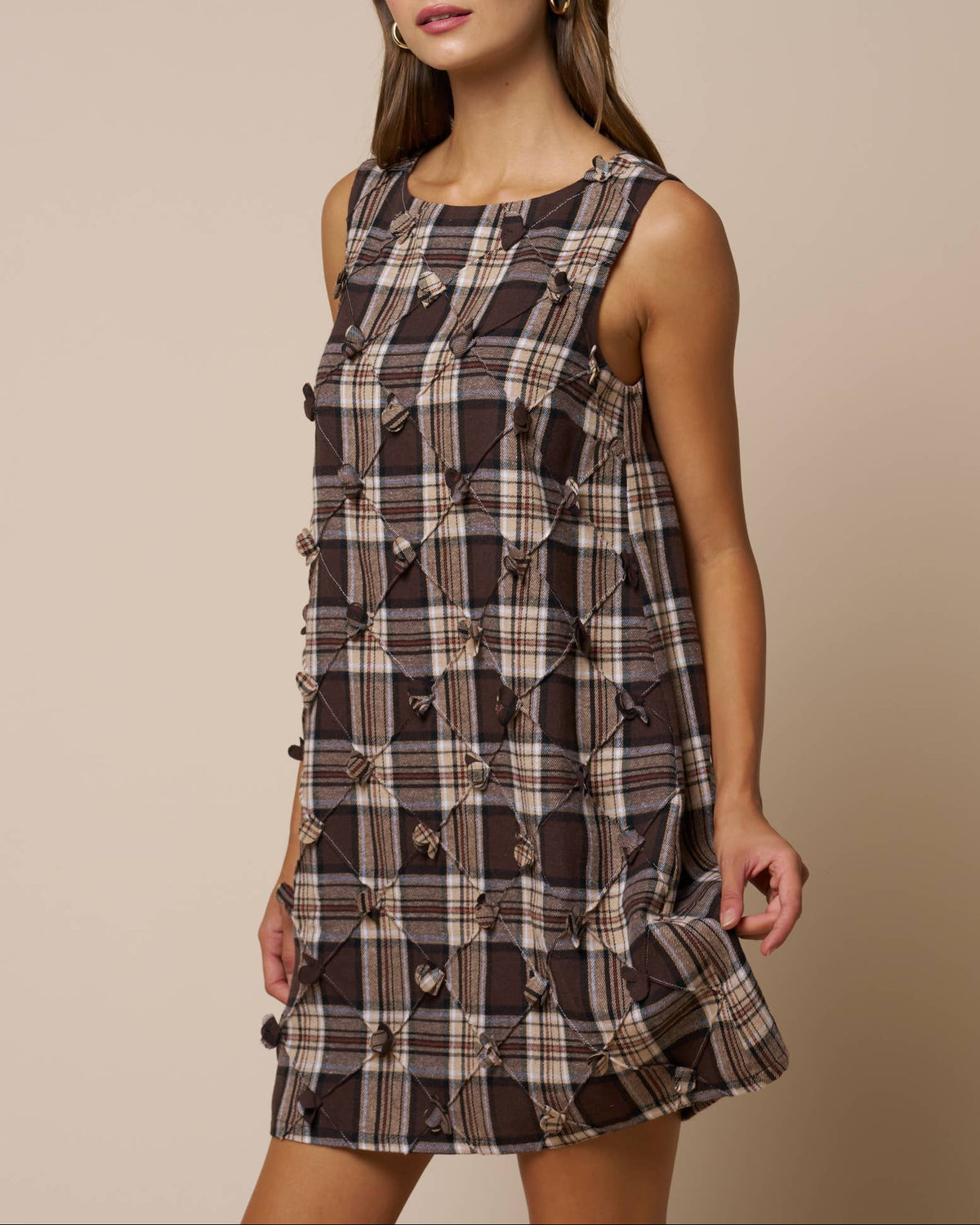 Model weaing a brown/taupe plaid sleeveless mini dress with diamond flower details on a neutral background side view