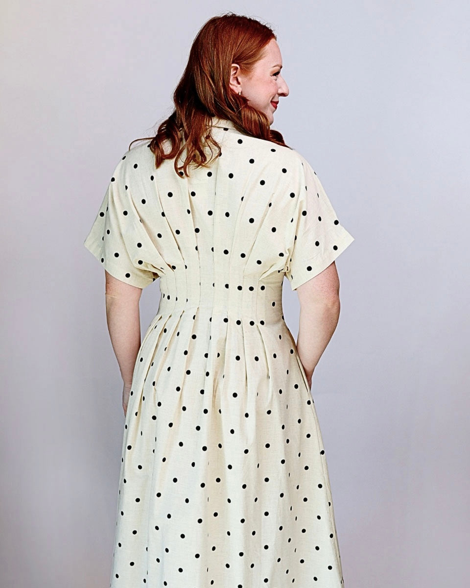 A woman with red hair is shown from the back wearing a cream-colored dress with black polka dots. The dress features short sleeves, a buttoned collar, and a matching fabric belt at the waist.