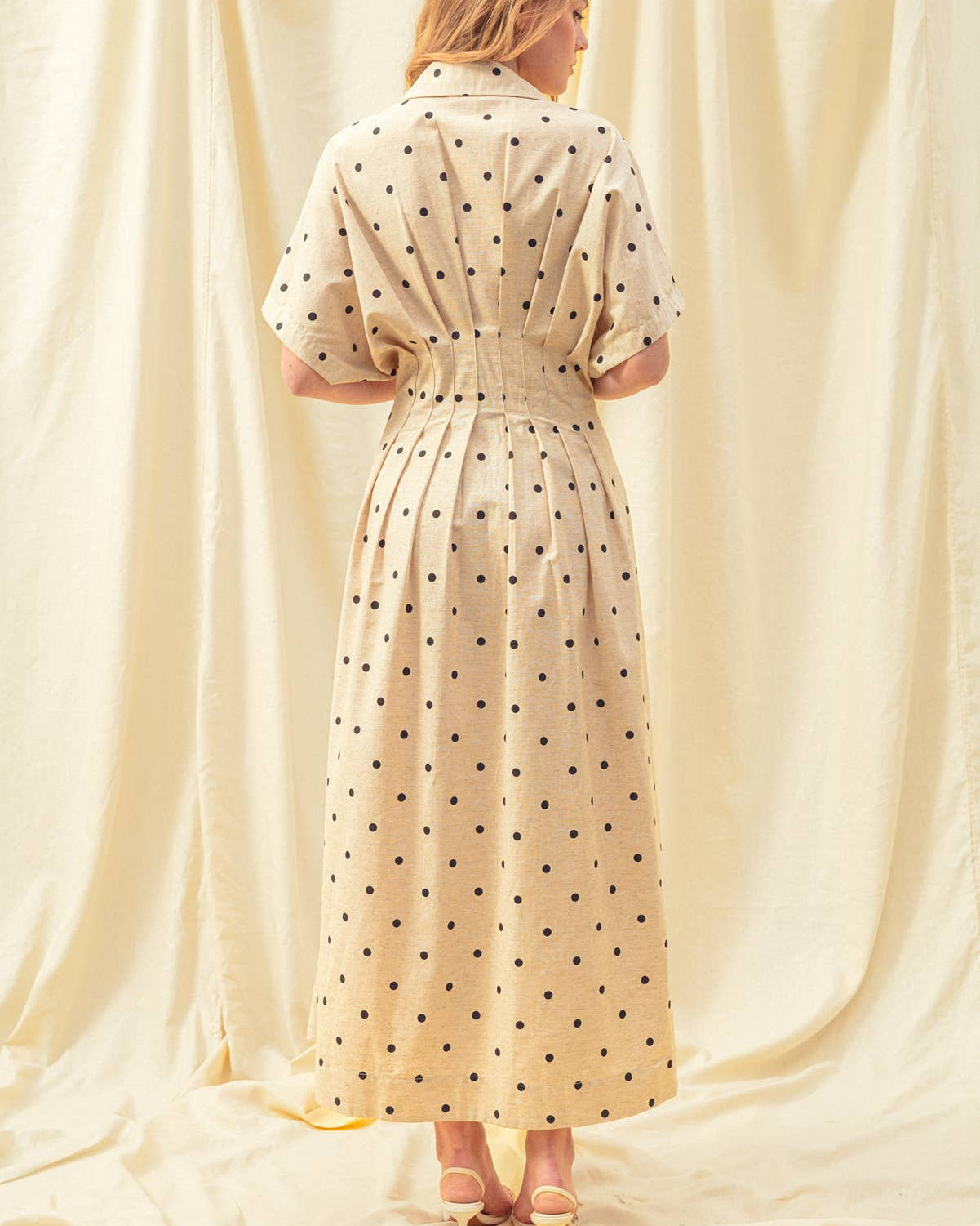 Woman wearing a beige dress with black polka dots in front of white curtains