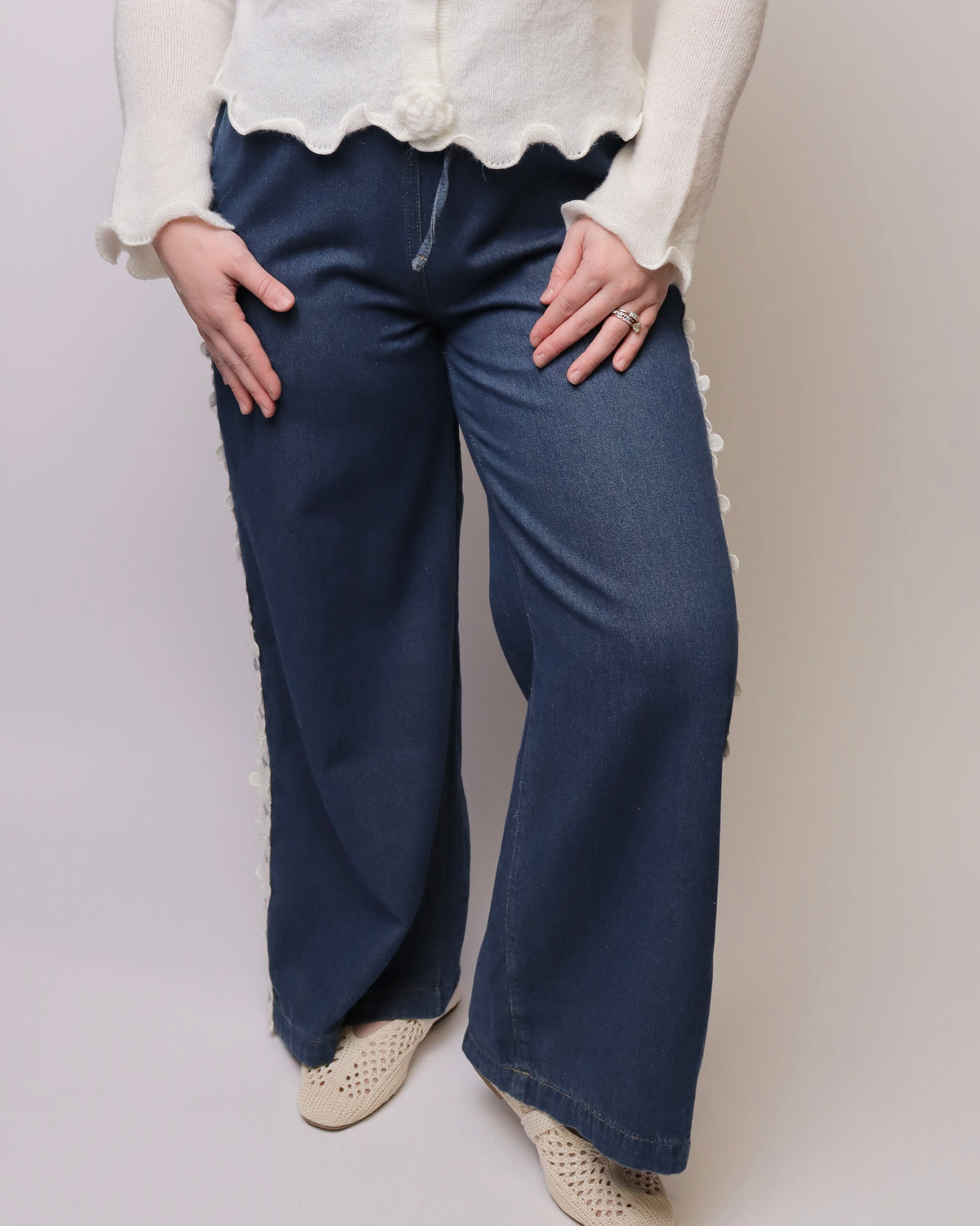A person wearing dark blue wide-leg jeans with white decorative pom-pom trim along the outer seams, paired with a cream knit top and beige perforated shoes, against a light gray studio background.
