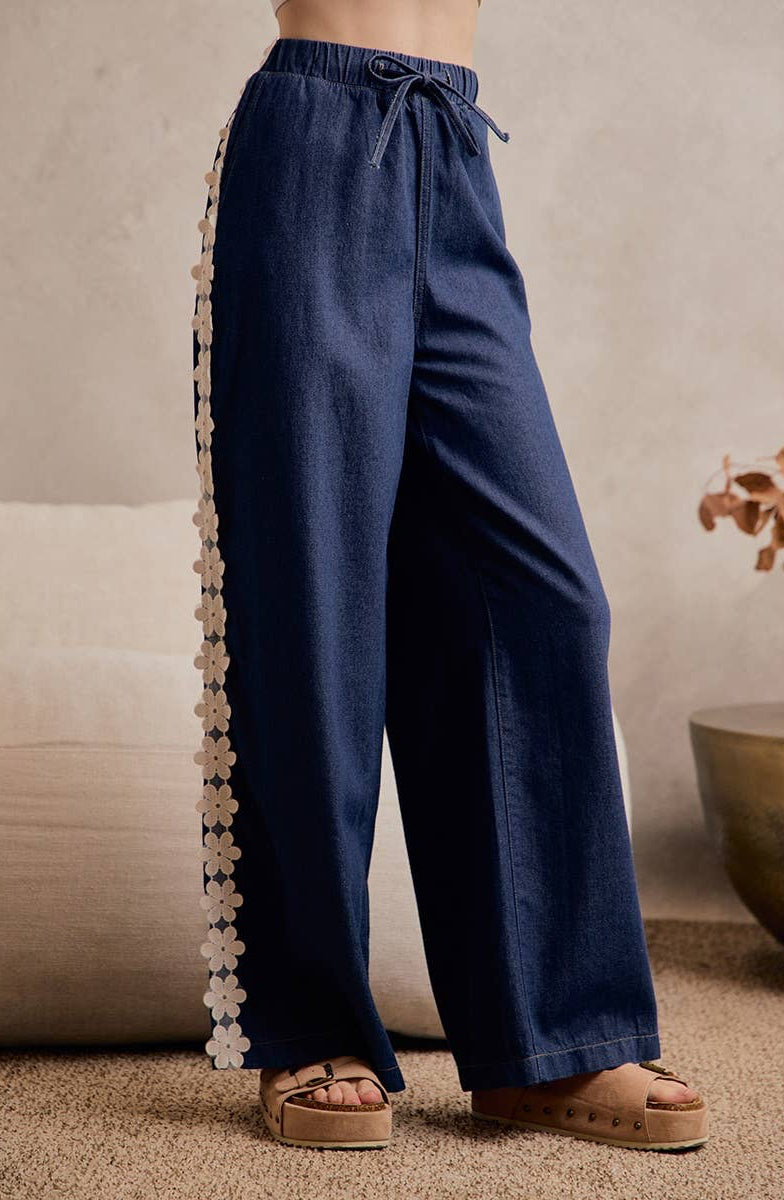 A person wearing dark blue wide-leg pants with a beige floral lace detail down the side, paired with tan platform sandals in a styled indoor setting.