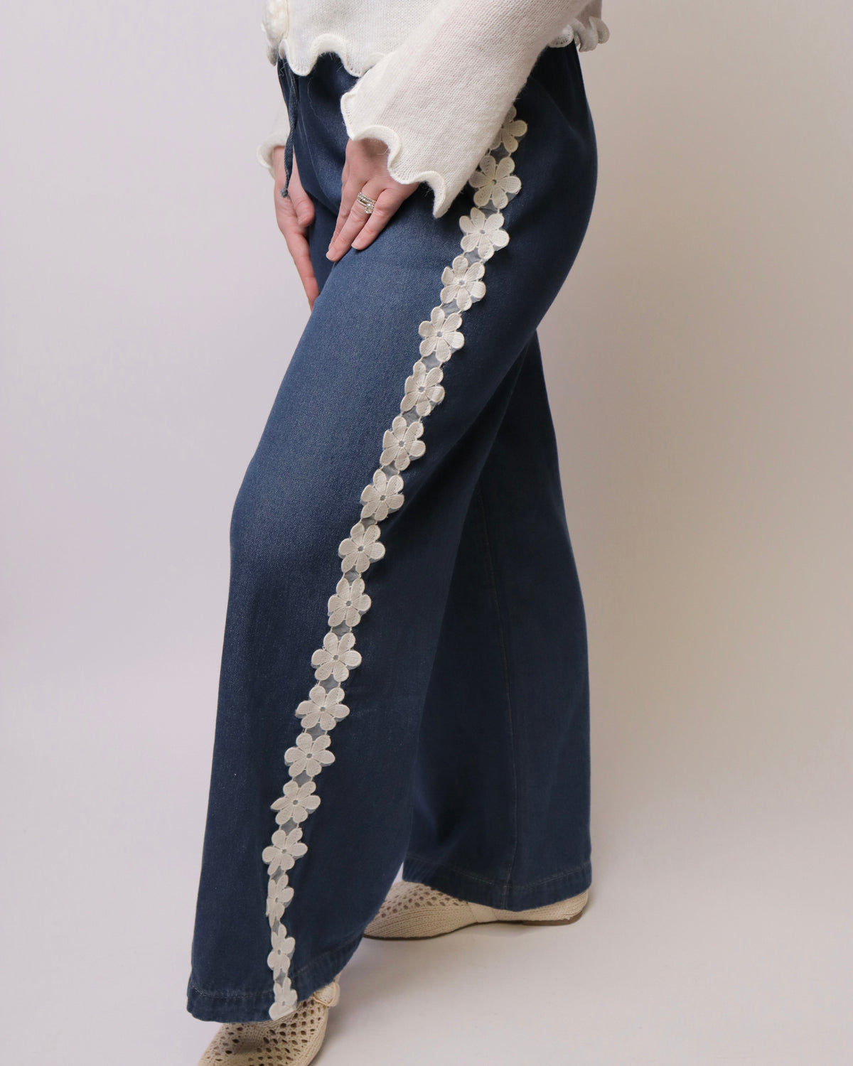 A person wearing dark blue wide-leg jeans with a vertical white floral lace applique down the side, paired with a white ruffled sweater and beige shoes, photographed against a plain light background.
