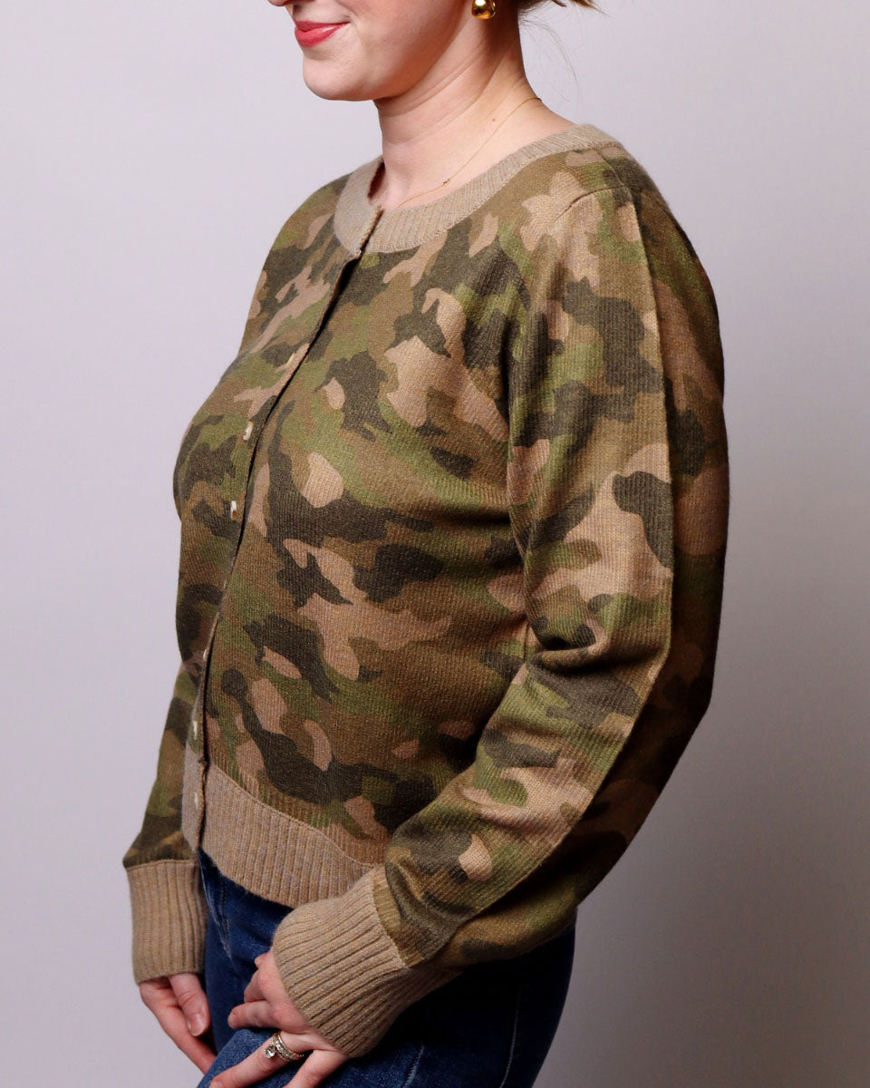 A woman wearing a long-sleeve knit cardigan with an olive green and tan camouflage pattern, paired with blue jeans, shown from a side profile against a plain light gray background.