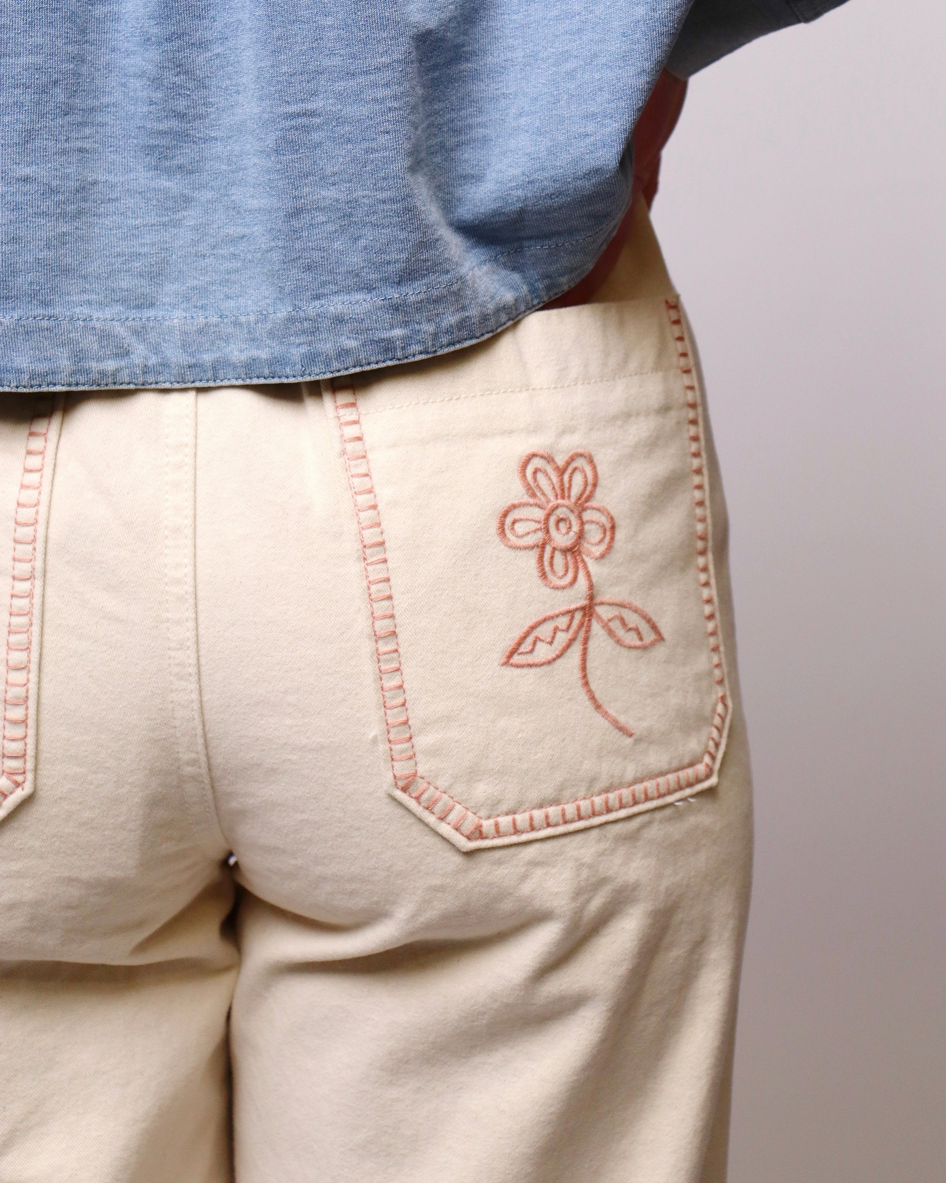 Close-up of a person wearing cream-colored pants with a pink embroidered flower on the back pocket, paired with a light blue denim shirt.