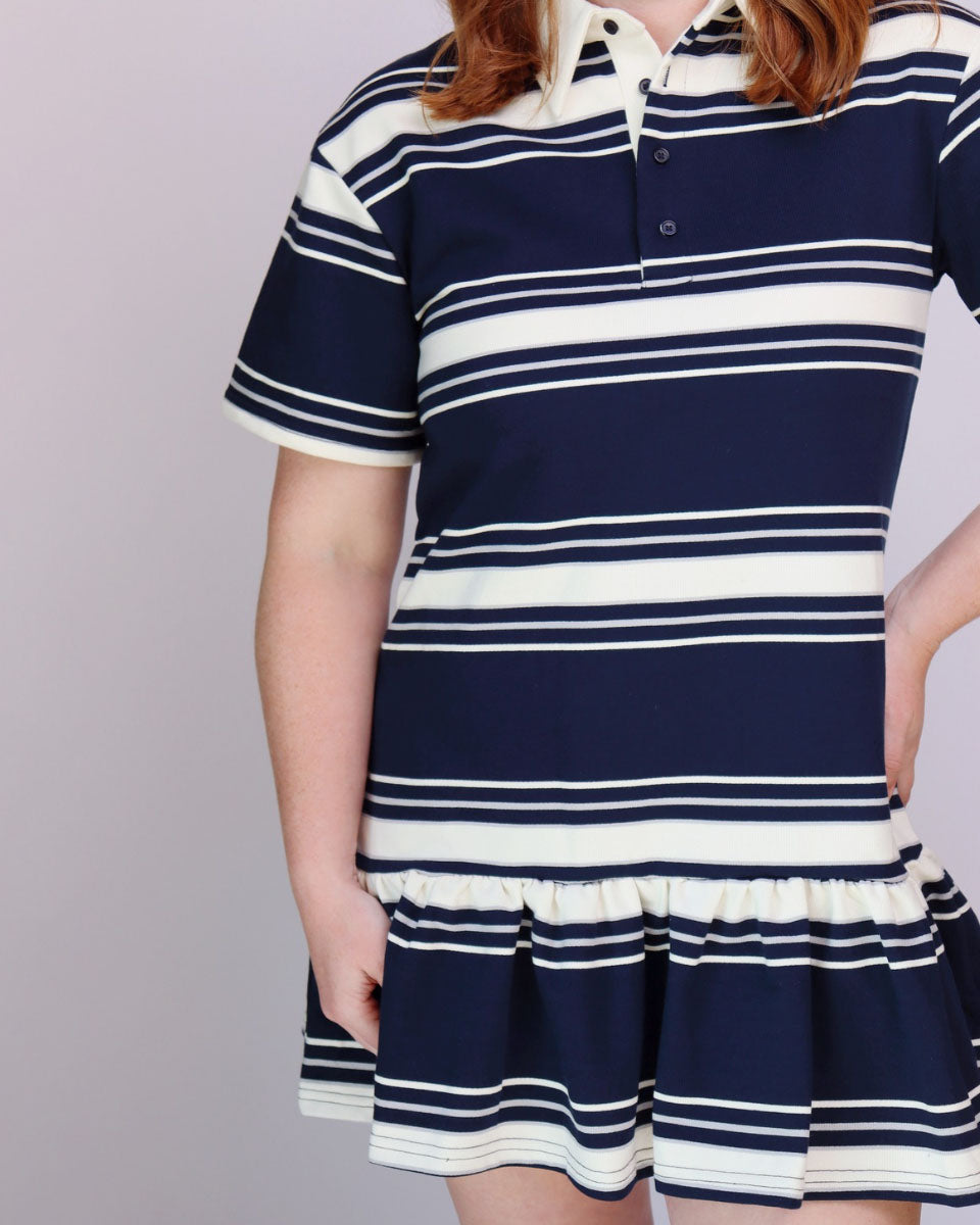 A child wearing a short-sleeve polo dress with horizontal navy blue and white stripes, featuring a collar, button placket, and a ruffled hem, shown against a plain light gray studio background.