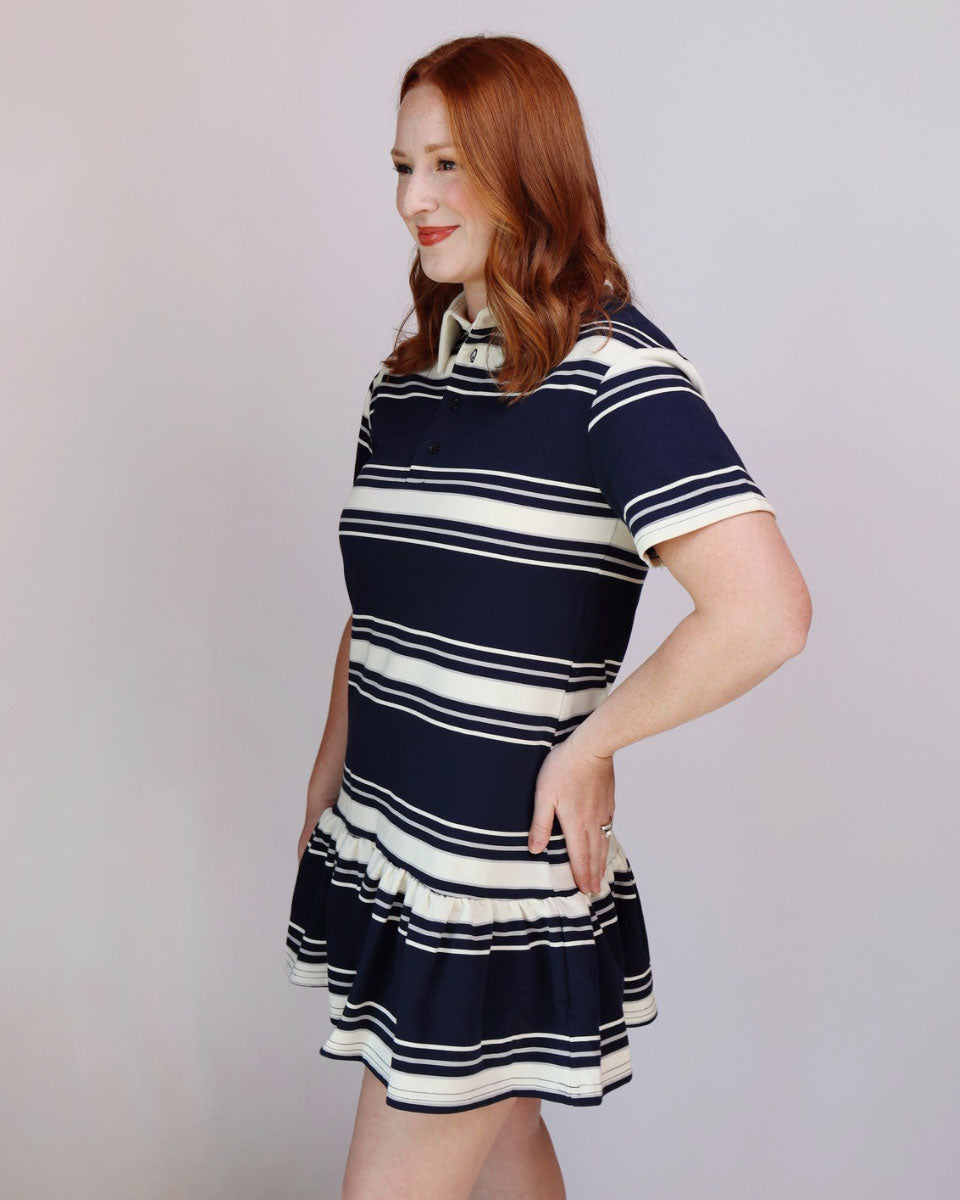 A woman with red hair wearing a short-sleeve polo-style mini dress with horizontal navy blue and cream stripes, standing against a plain light gray studio background.