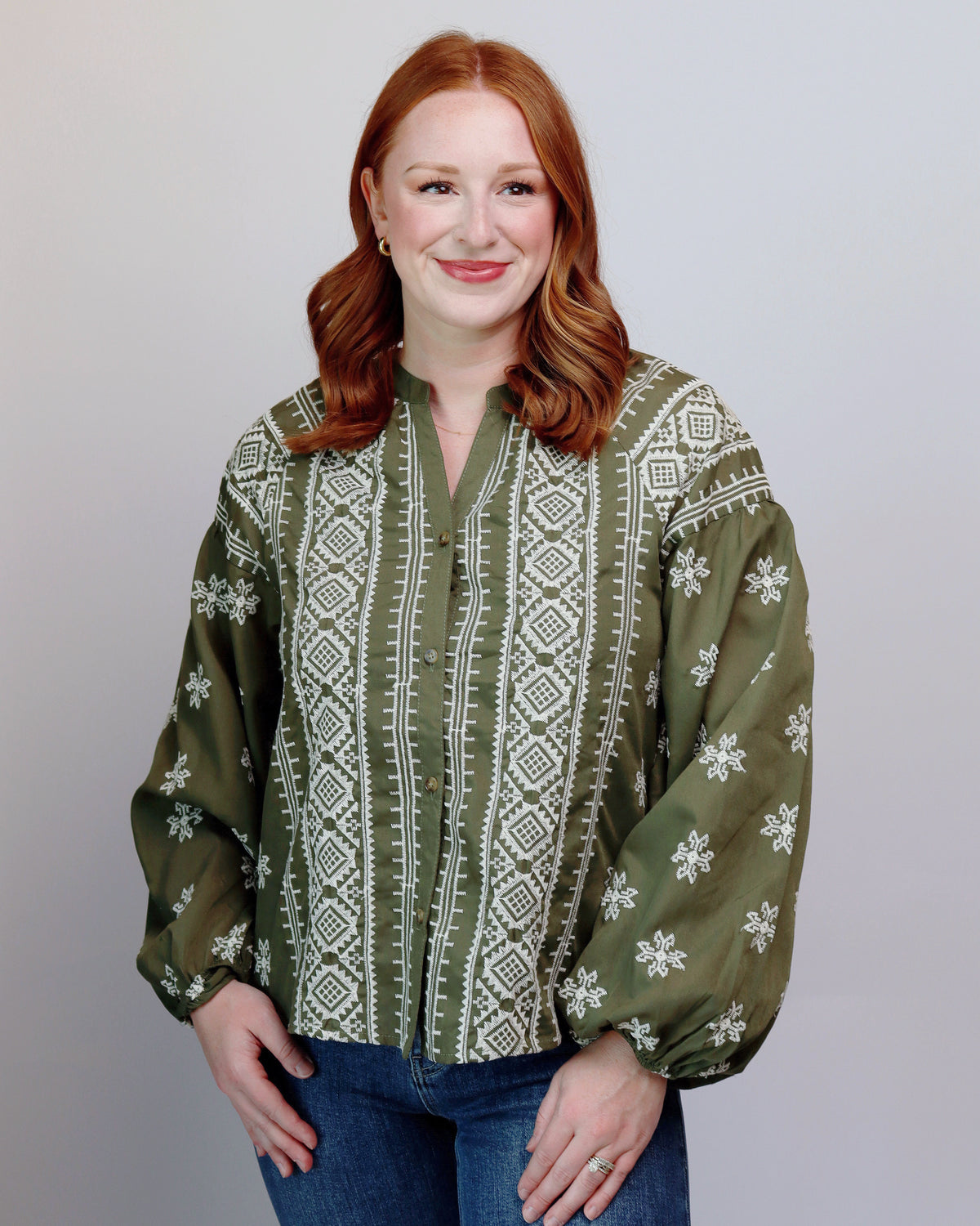 A woman with red hair wearing a green long-sleeve blouse with white geometric and snowflake patterns, paired with blue jeans, standing against a light gray background.