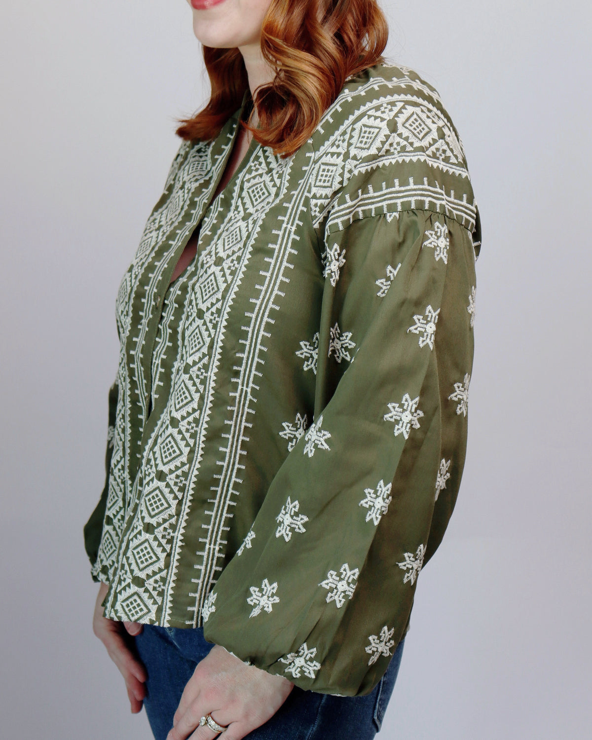 A woman wearing an olive green long-sleeve blouse with white geometric and floral embroidery, paired with blue jeans, shown against a plain light gray background.