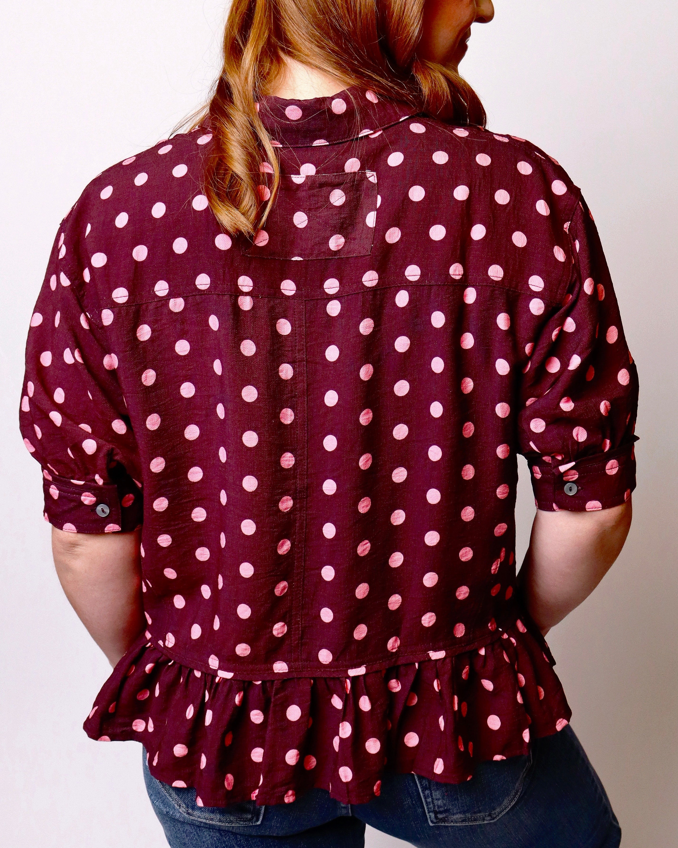 A woman shown from the back wearing a burgundy button-up blouse with pink polka dots, rolled sleeves, and a ruffled hem, paired with blue jeans against a plain light background.