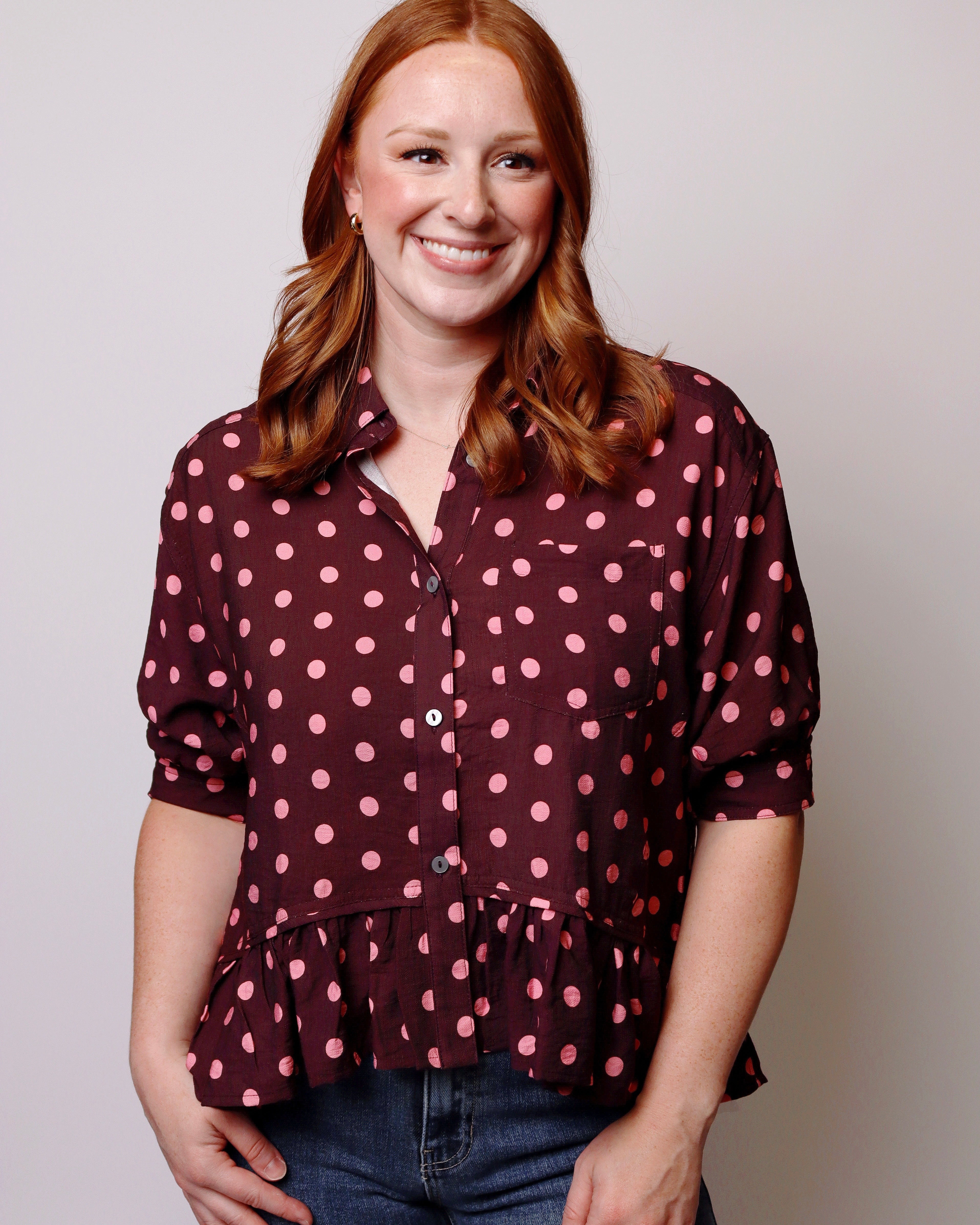 A smiling woman with red hair wearing a dark burgundy button-up blouse with pink polka dots and rolled sleeves, paired with blue jeans, against a plain light gray background.