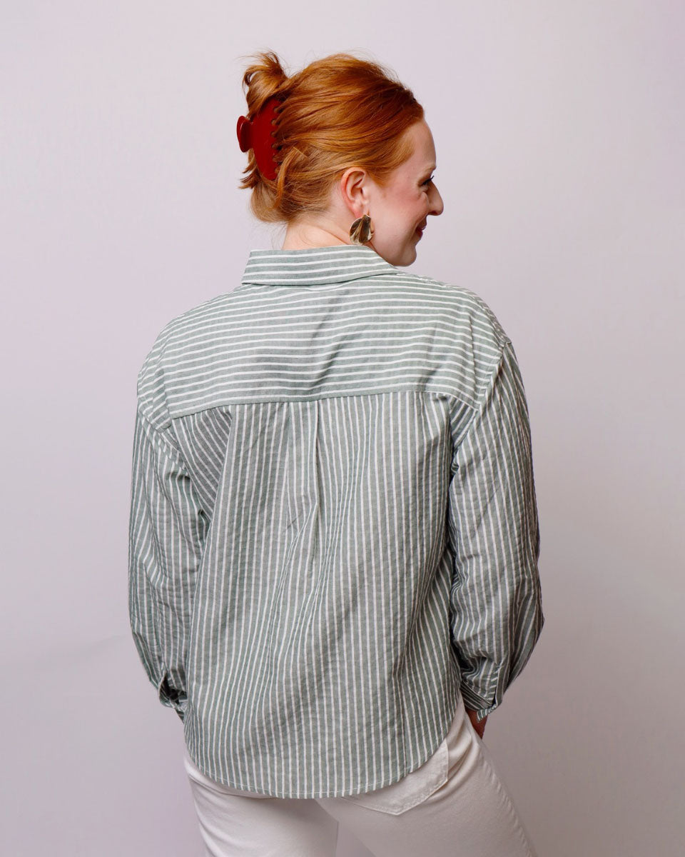 A woman with red hair tied up in a bun, viewed from behind, wearing a sage green and white vertically striped button-down shirt and white pants against a light gray background.