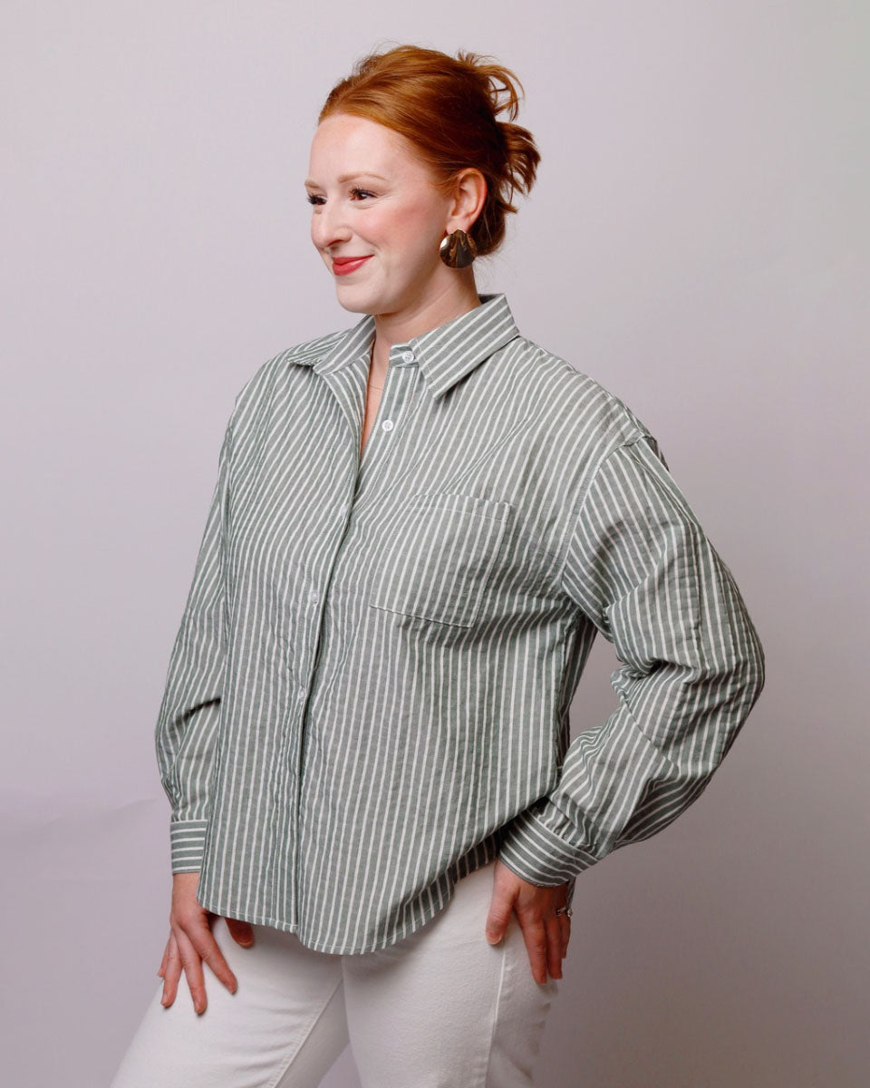 A woman with red hair tied back is wearing a light sage green and white vertically striped button-up shirt with white pants, posing against a solid light gray studio background.
