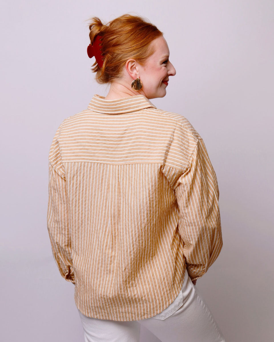 A woman with red hair tied up in a bun is shown from the back wearing a beige and white vertically striped button-up shirt and white pants, set against a plain light gray studio background.