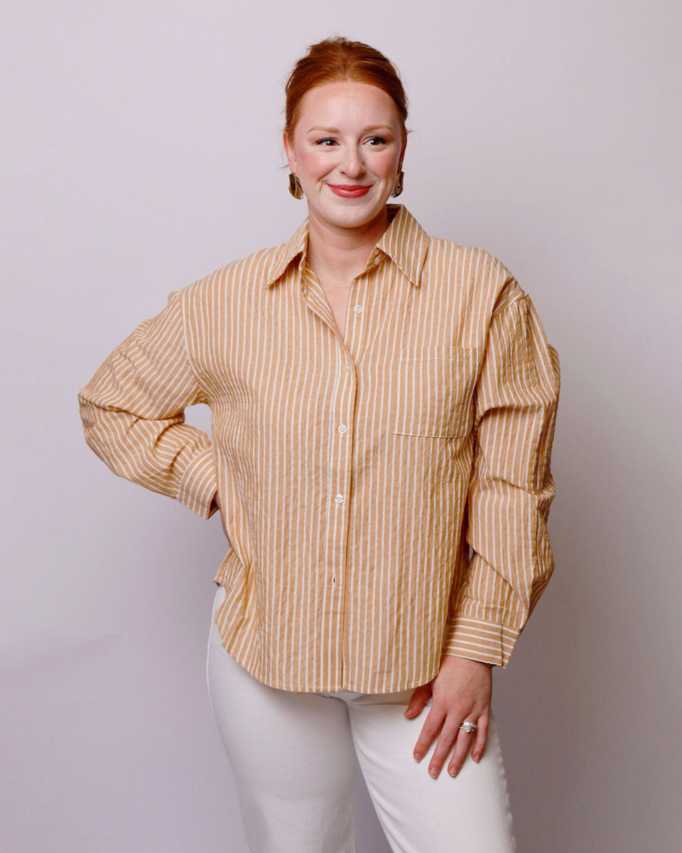 A woman with red hair tied back, wearing a beige and white vertically striped button-down shirt and white pants, posing against a light gray studio background.