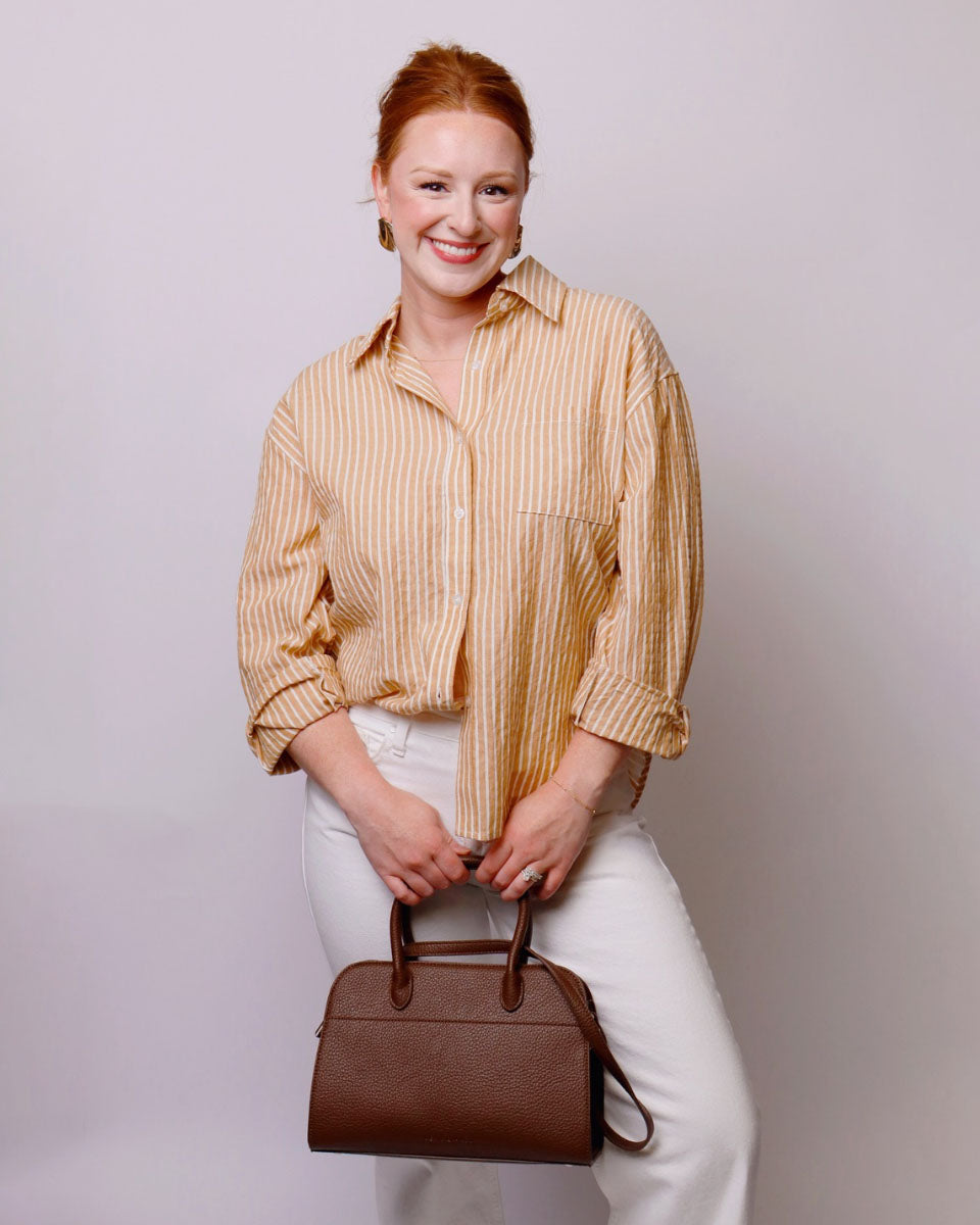 A smiling woman with red hair stands against a light gray studio background, wearing a beige and yellow vertically striped button-up shirt, white pants, and holding a brown leather handbag.