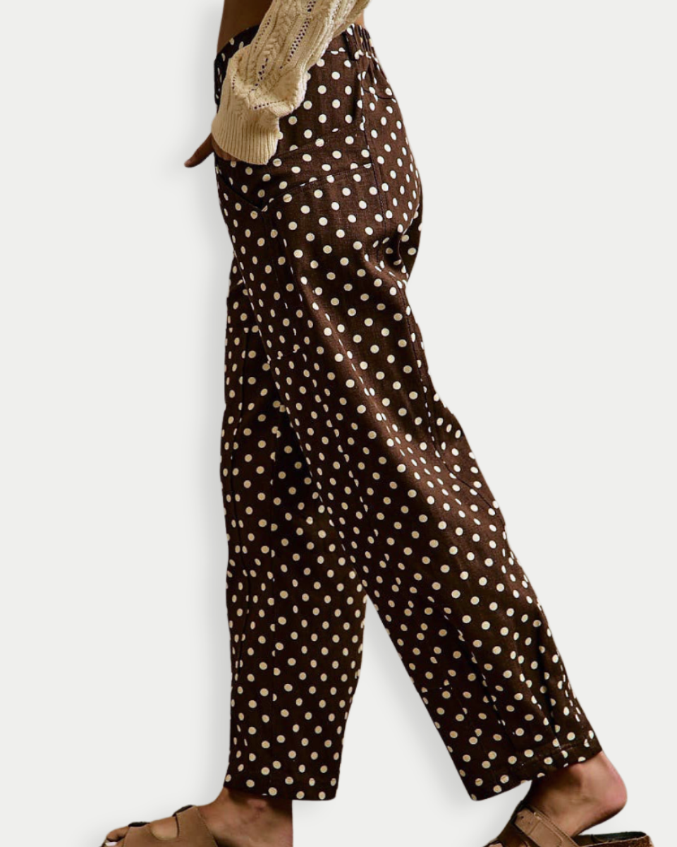A side view of a person wearing brown wide-leg trousers with a white polka dot pattern, paired with a beige knit sweater and brown slide sandals, against a light gray background.