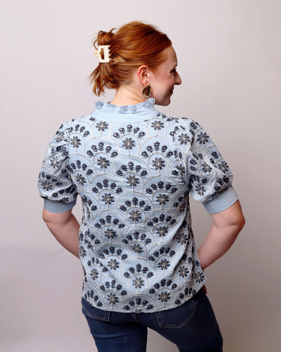 A woman with red hair tied back in a bun is shown from the back, wearing a light blue short-sleeve blouse with a dark blue floral pattern, paired with dark blue jeans against a plain light gray background.