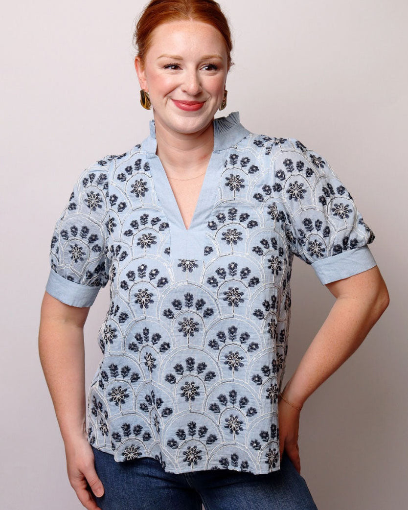 A smiling woman with red hair wearing a light blue short-sleeve blouse with a dark blue floral pattern, paired with blue jeans, against a plain light gray background.