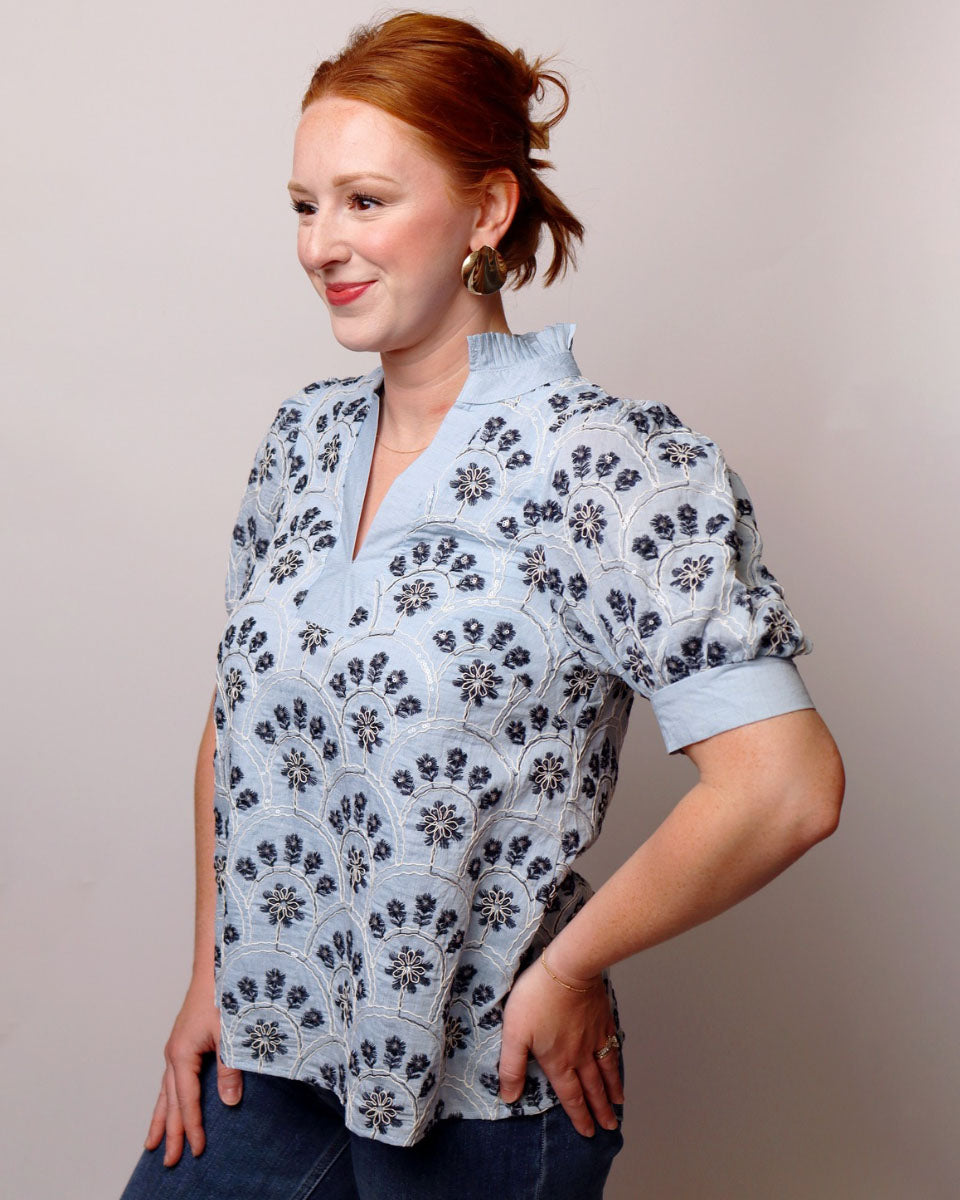 A woman with red hair tied back wears a light blue short-sleeve blouse with a dark blue floral pattern, paired with dark blue jeans, against a plain light gray studio background.