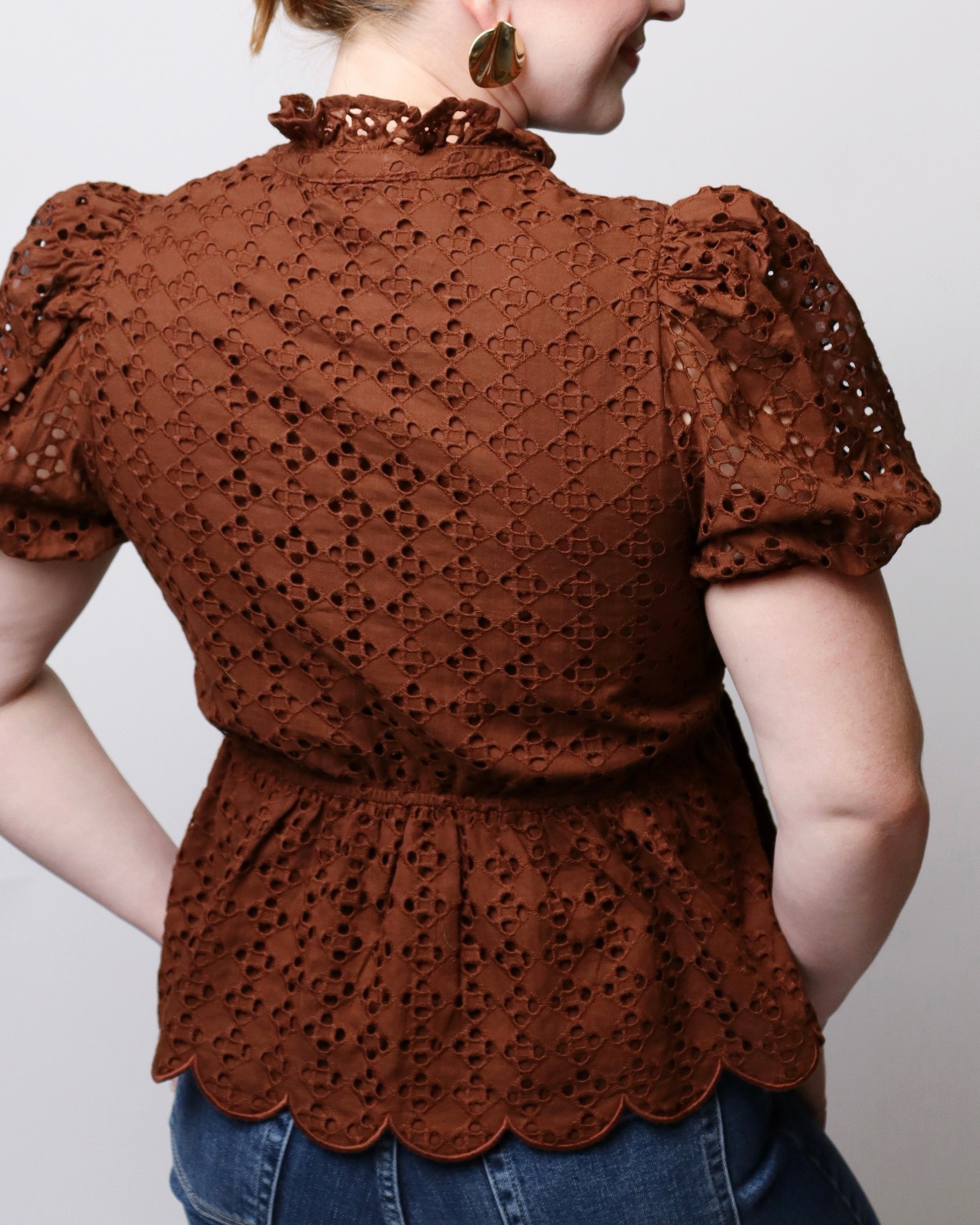 A woman wearing a brown eyelet lace blouse with puff sleeves, a ruffled high neck, and a scalloped hem, shown from the back against a light gray background.
