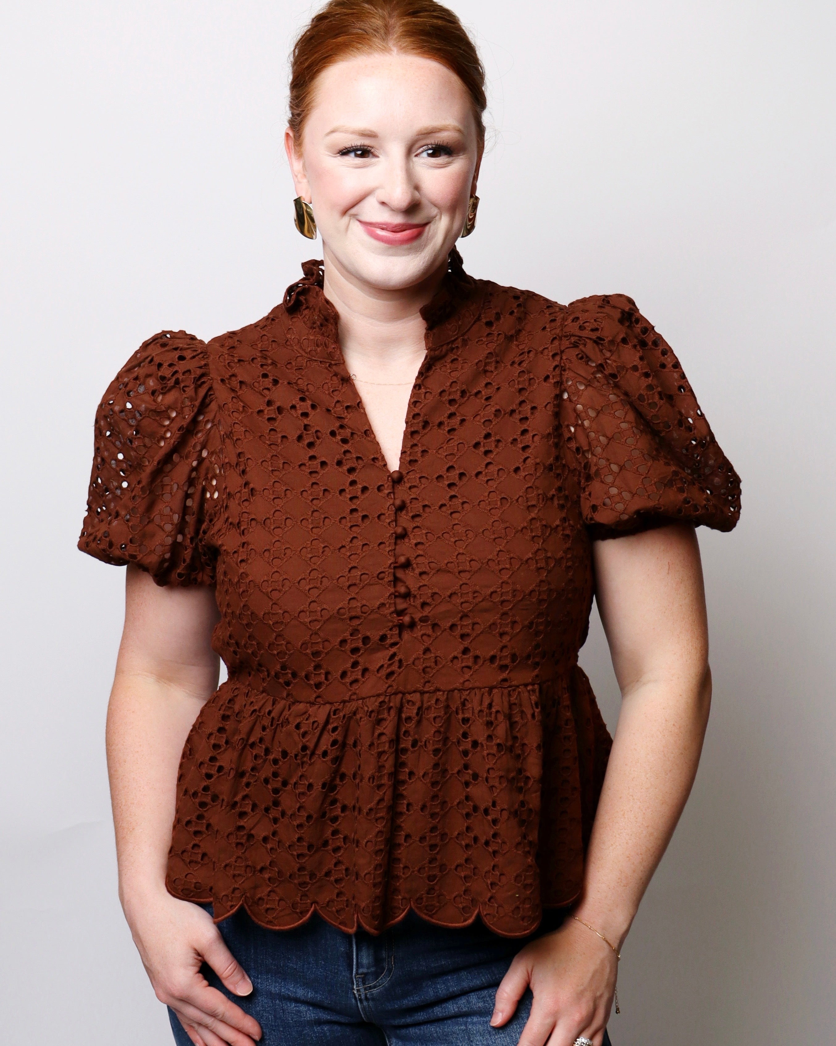 A woman modeling a brown eyelet lace peplum top with puff sleeves and a scalloped hem, paired with blue jeans, against a light gray studio background.