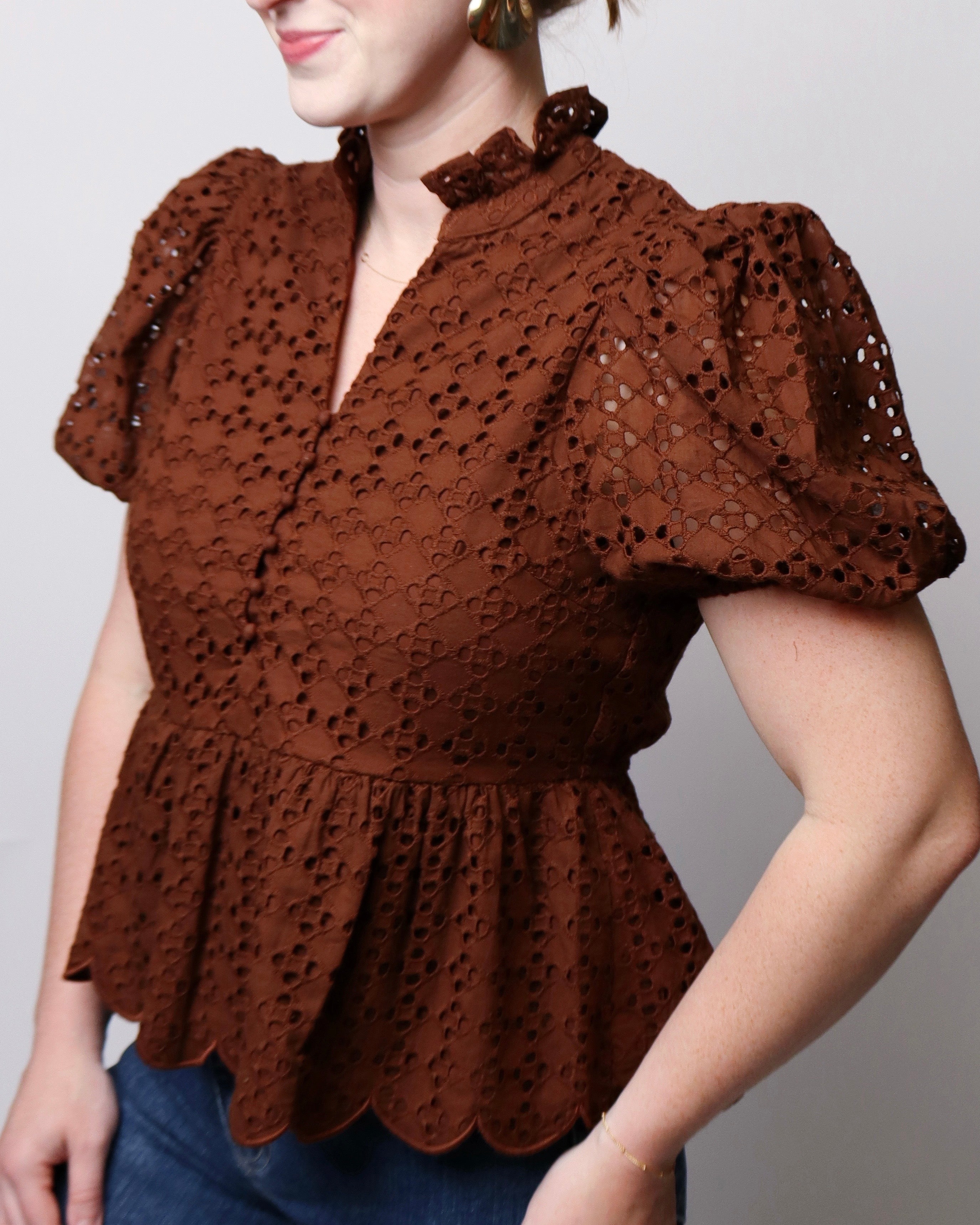 A woman wearing a brown eyelet lace blouse with puff sleeves, a ruffled high neckline, and a scalloped peplum hem, paired with blue jeans against a plain light gray background.
