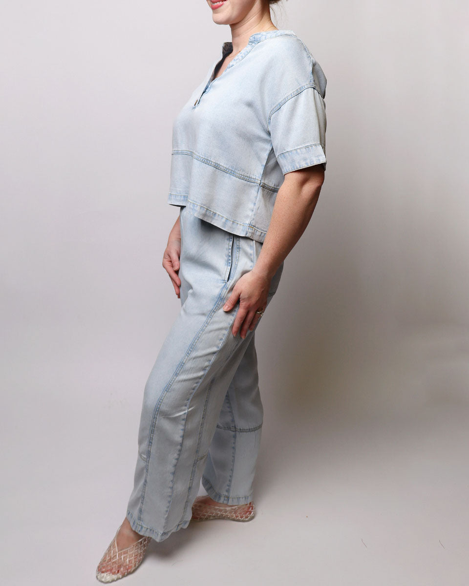 A woman modeling a light blue denim two-piece set consisting of a short-sleeve cropped top and matching wide-leg pants, photographed against a neutral light gray studio background.