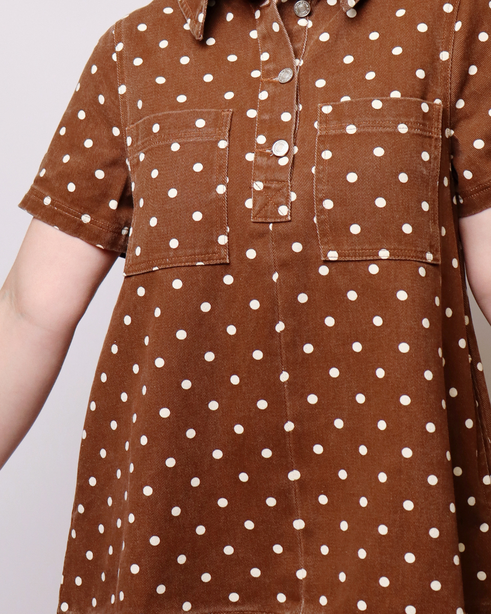 A close-up of a brown corduroy short-sleeve shirt dress with a white polka dot pattern, featuring a buttoned collar and two chest pockets.
