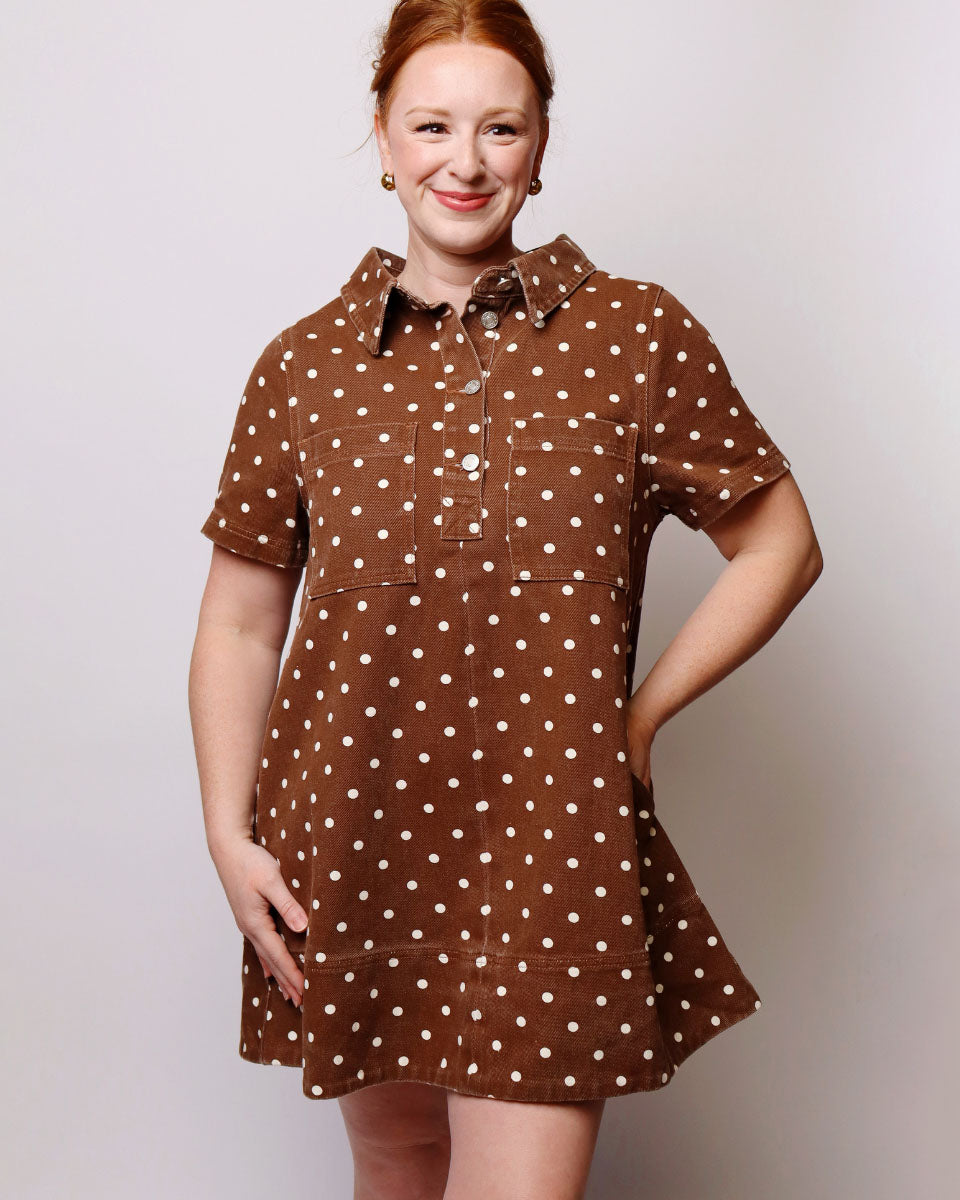 A woman modeling a brown short-sleeve collared dress with white polka dots, featuring a buttoned placket and chest pockets, set against a plain light gray background.