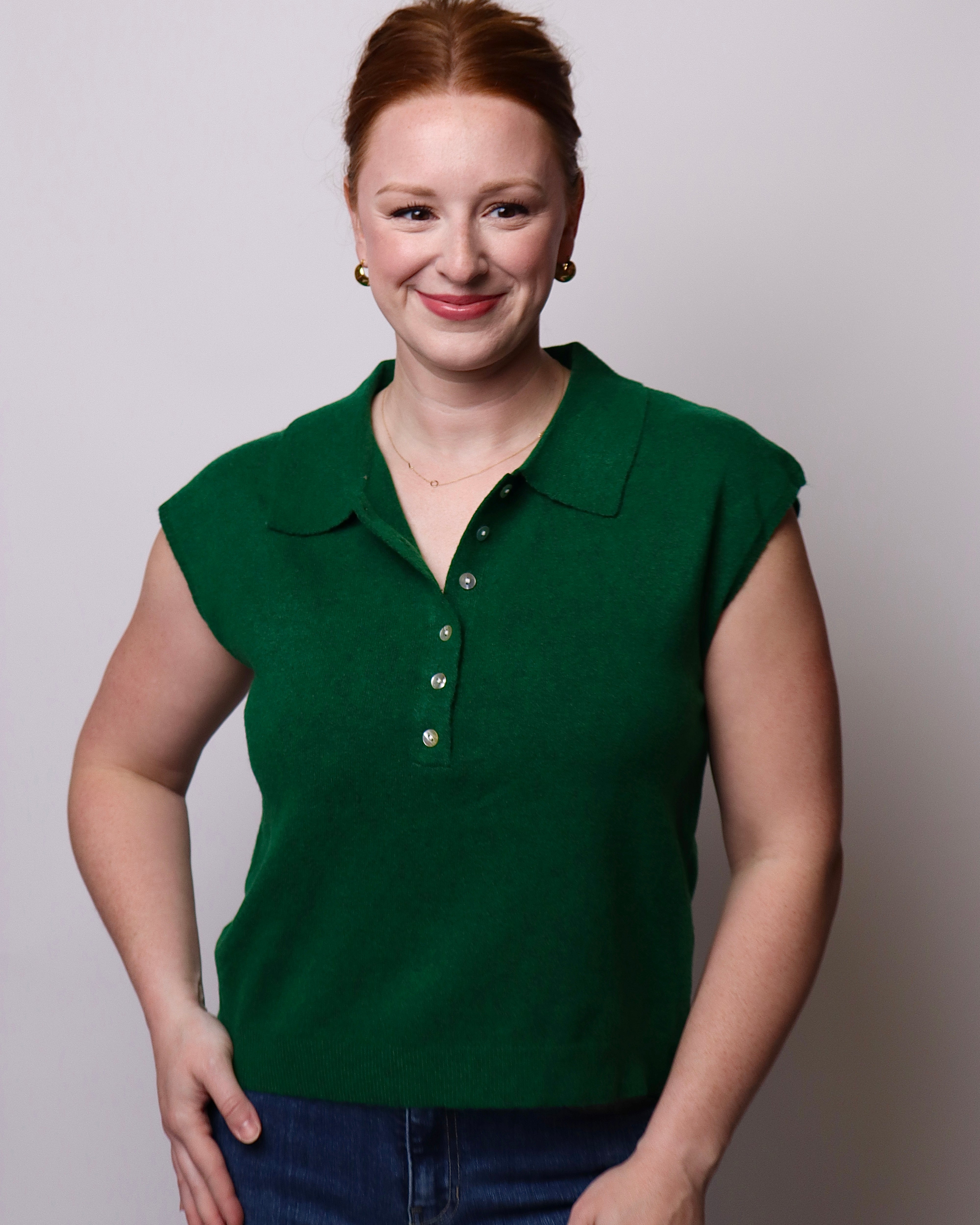 A woman with red hair tied back, wearing a dark green short-sleeve knit polo shirt with a button placket and blue jeans, standing against a light gray studio background.