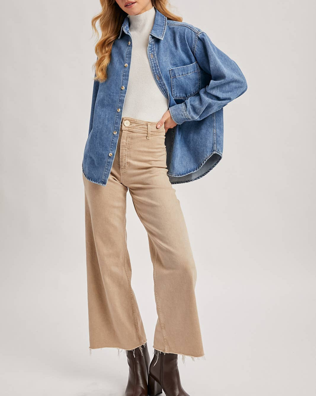Woman wearing a denim jacket, white turtleneck, beige pants, and brown boots on a light gray background